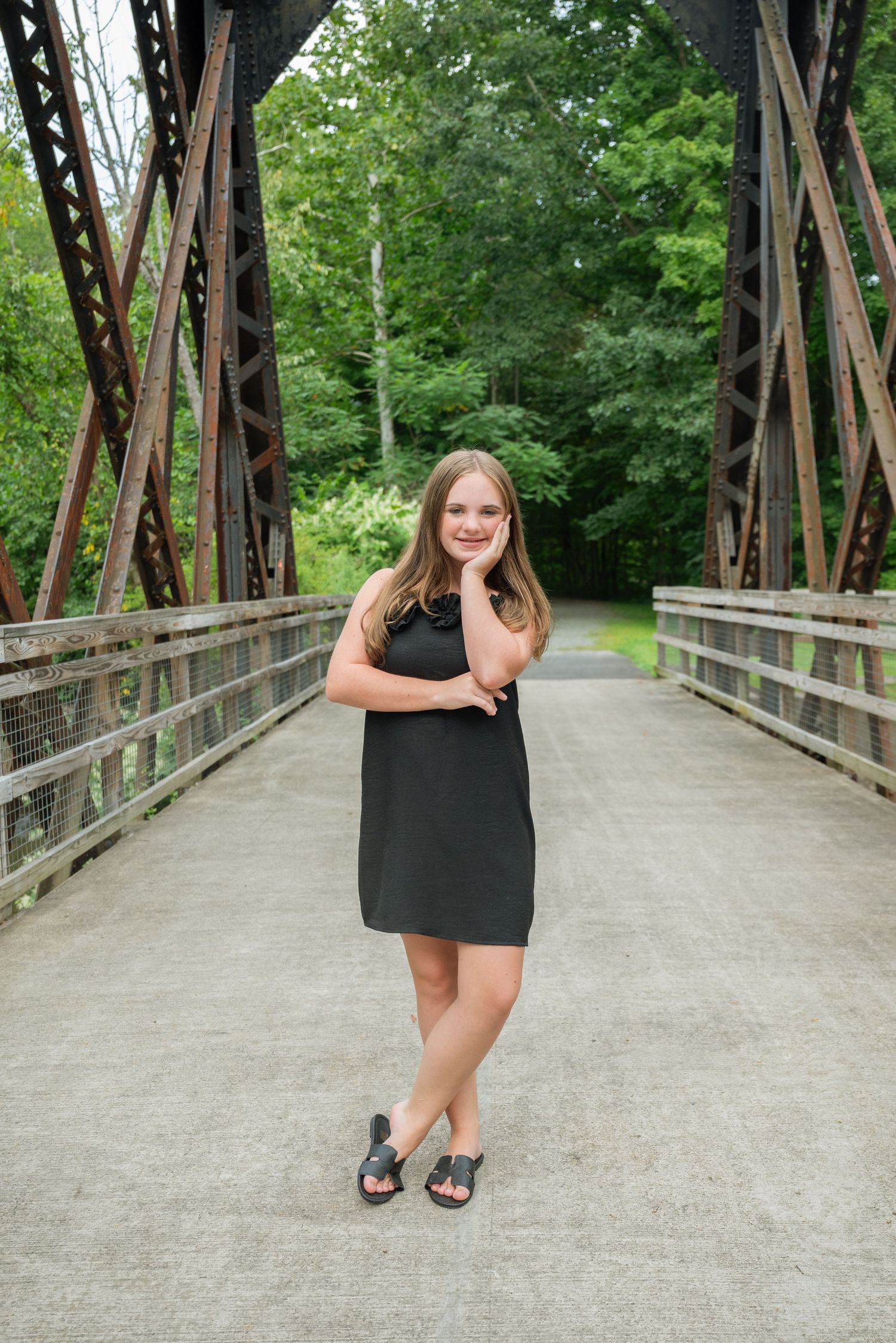 Young woman in black dress smiling on rustic iron bridge surrounded by lush green trees.