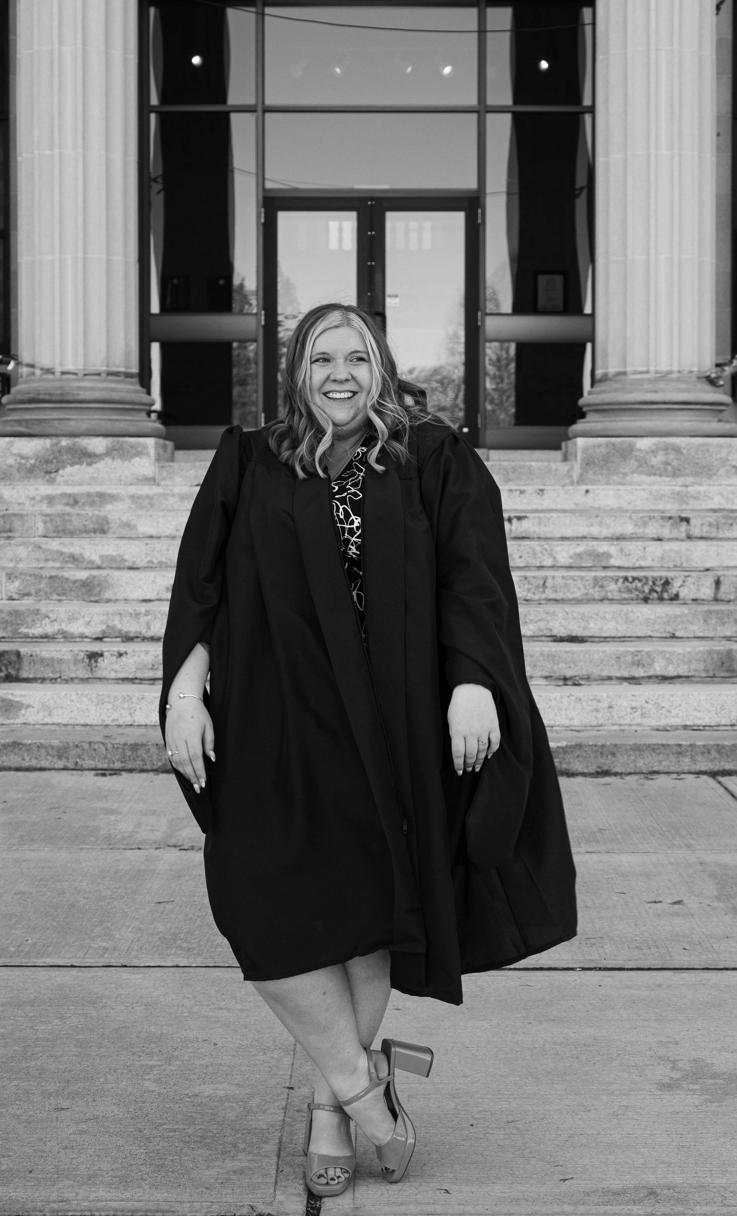 Smiling graduate in black gown standing before columned building entrance.