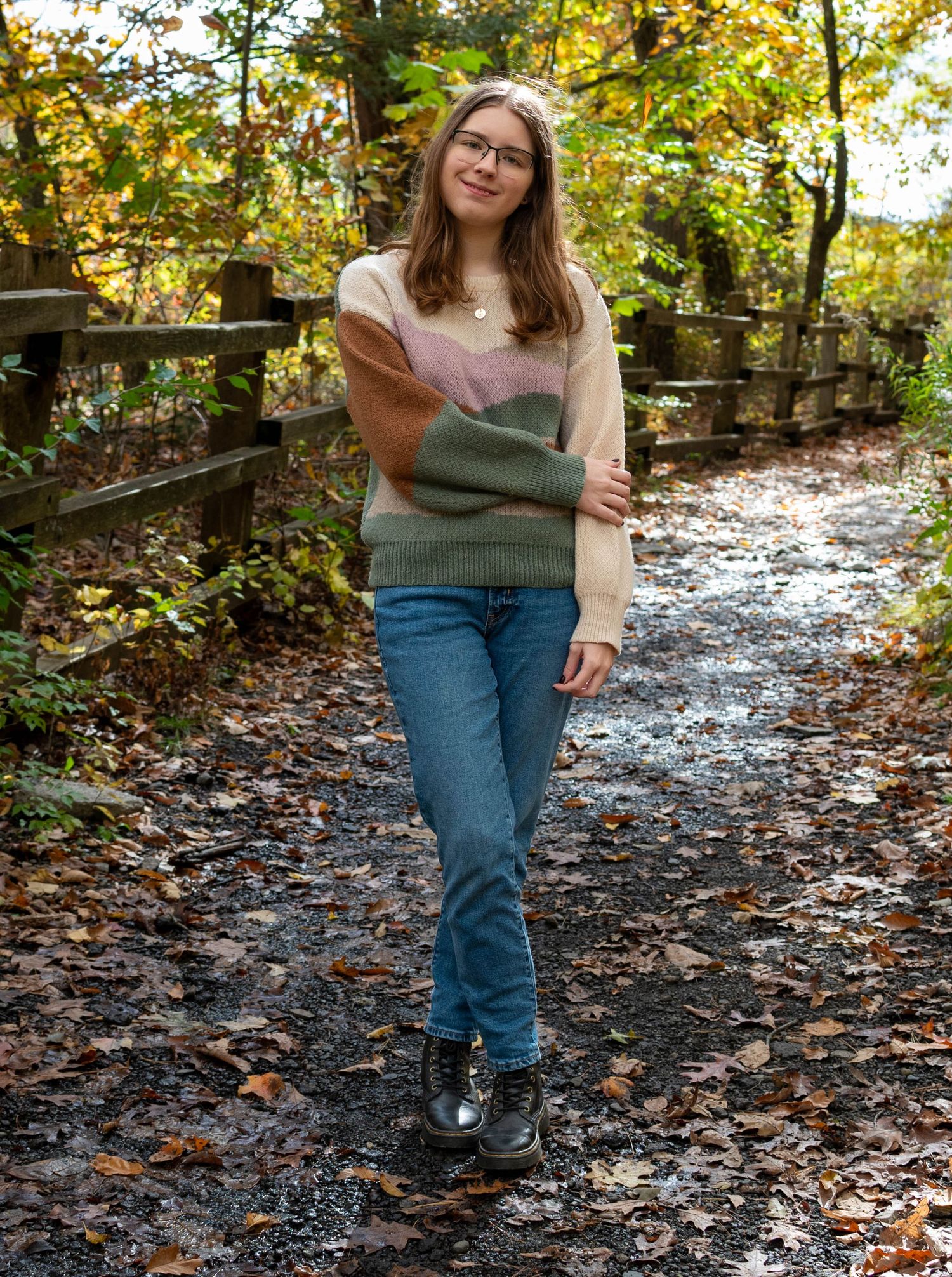 Teen girl in colorblock sweater and jeans walks autumn leaf-covered path beside wooden fence.