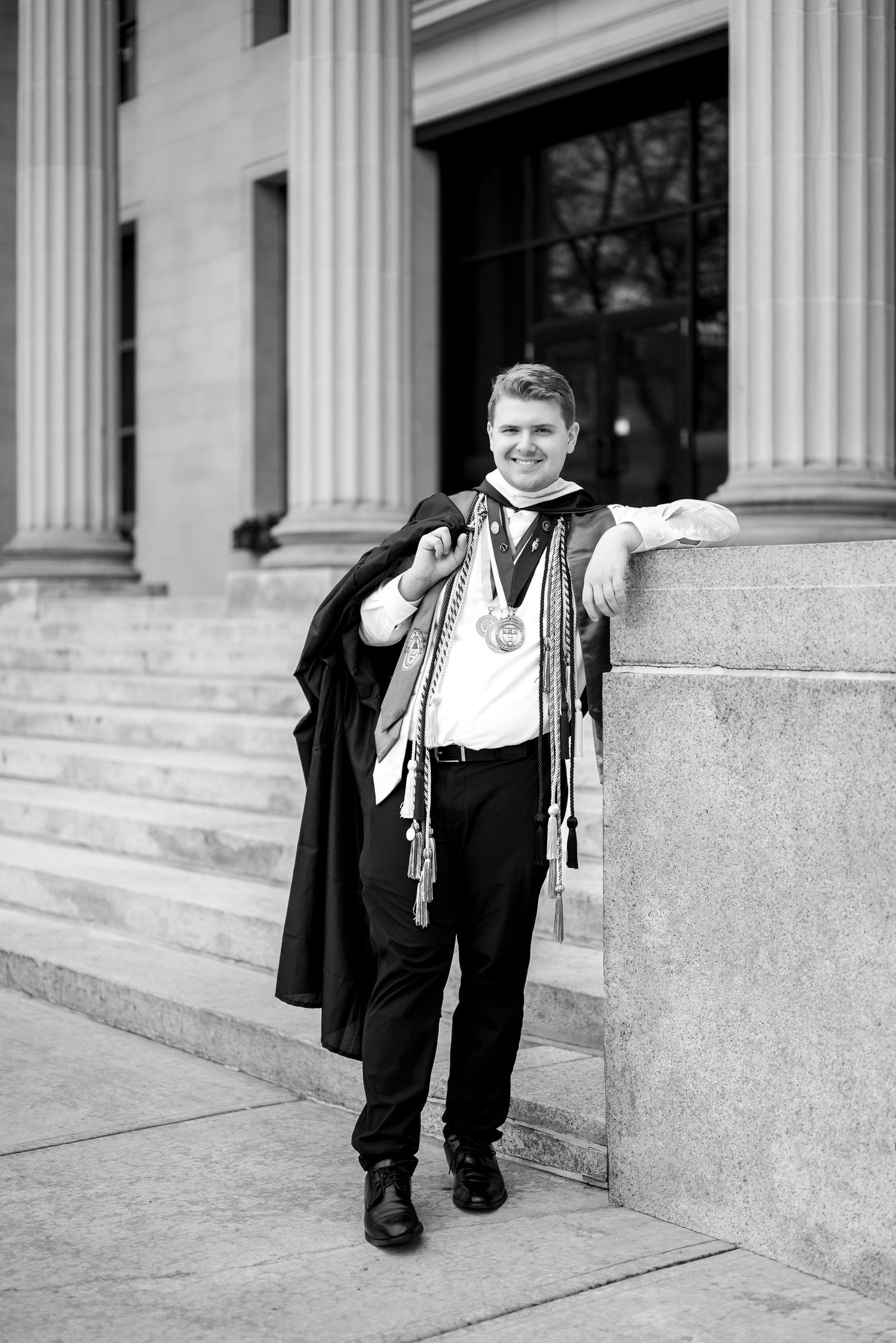 Black and white photo of a smiling graduate wearing medals and sashes, leaning against a stone column outside a university building.