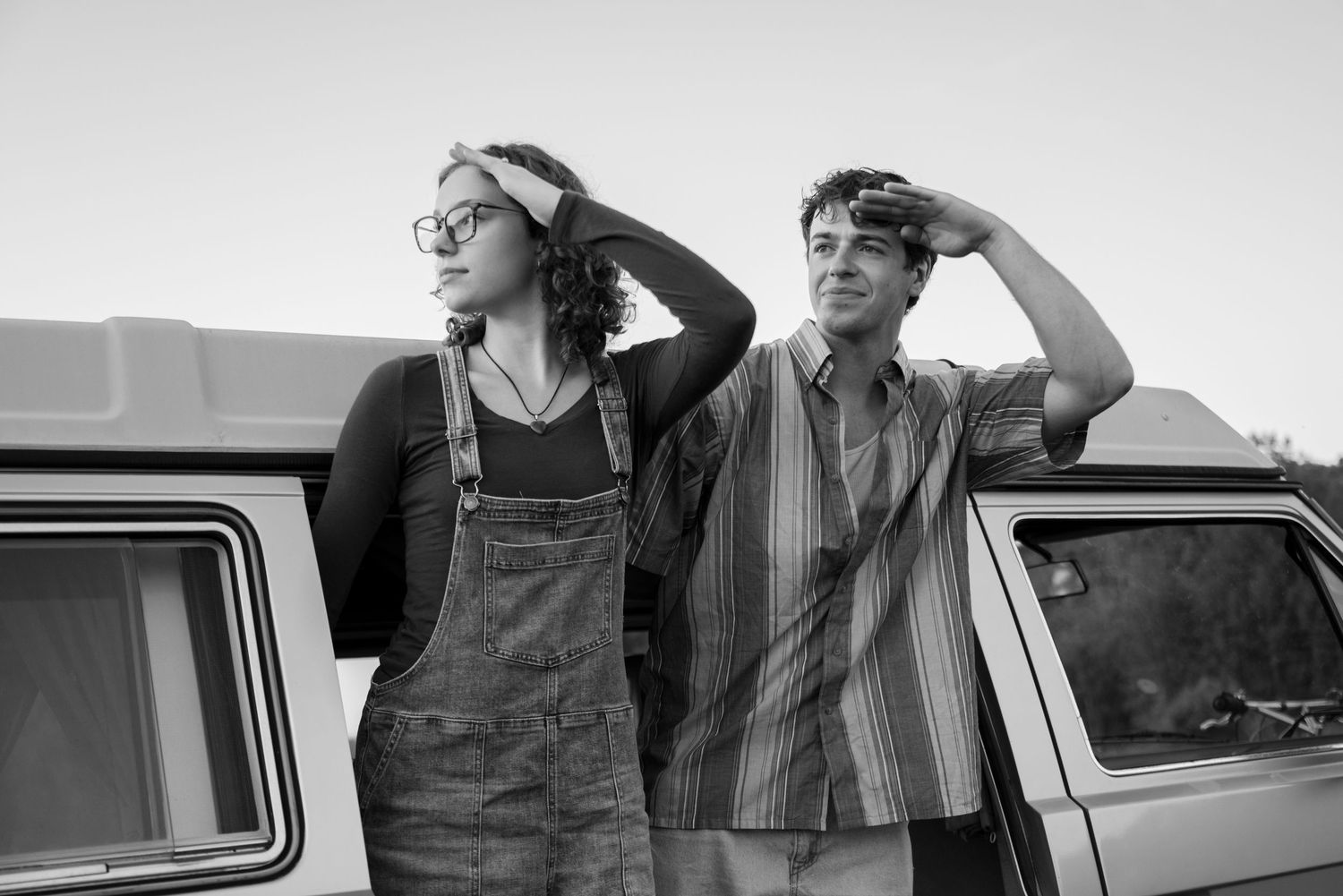 Two young people lean against a vintage van, shielding their eyes in a black and white retro-style outdoor photo.