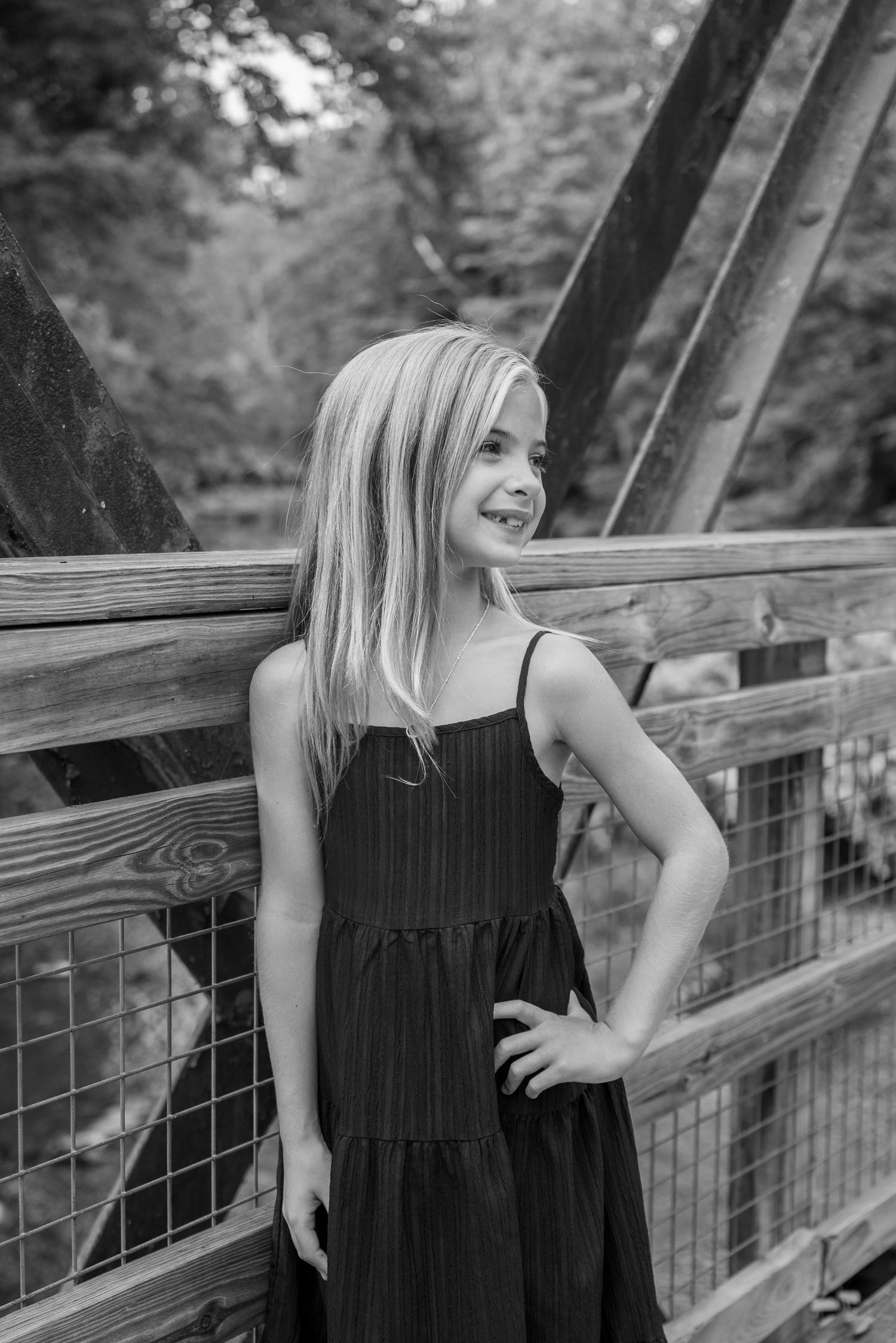 Black and white photo of a young blonde girl in a sundress posing on a wooden bridge outdoors.