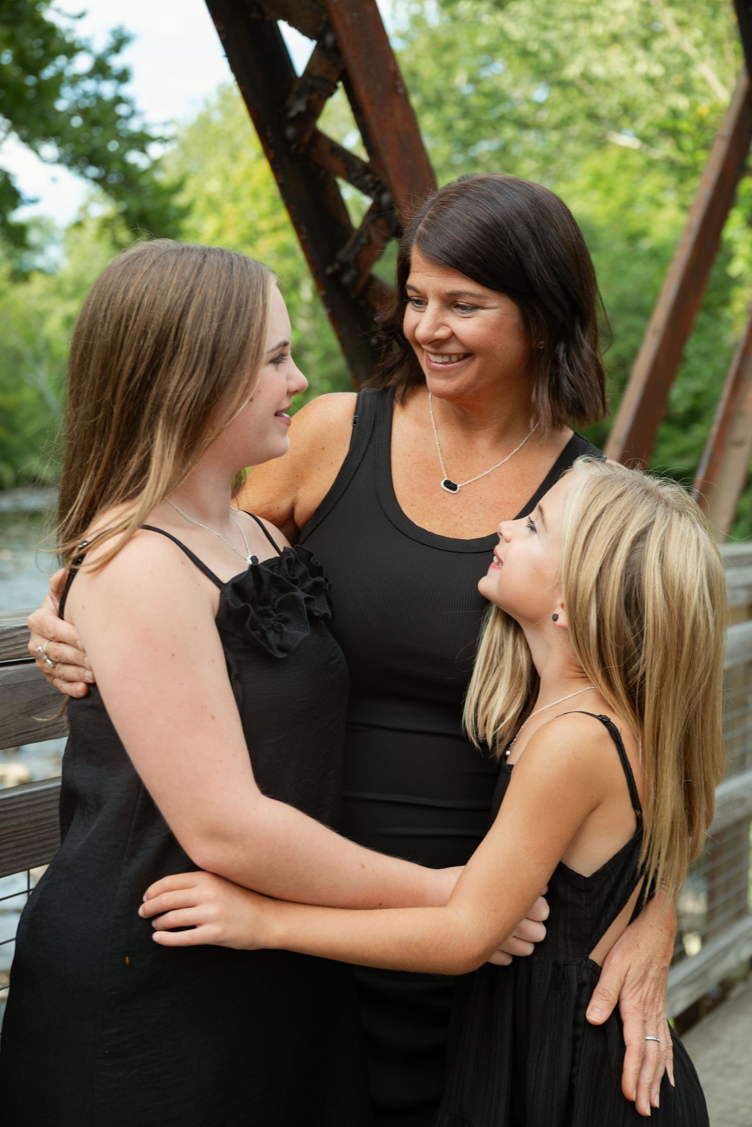 A smiling brunette mother embraces her two blonde daughters, all wearing black dresses, on a bridge outdoors.