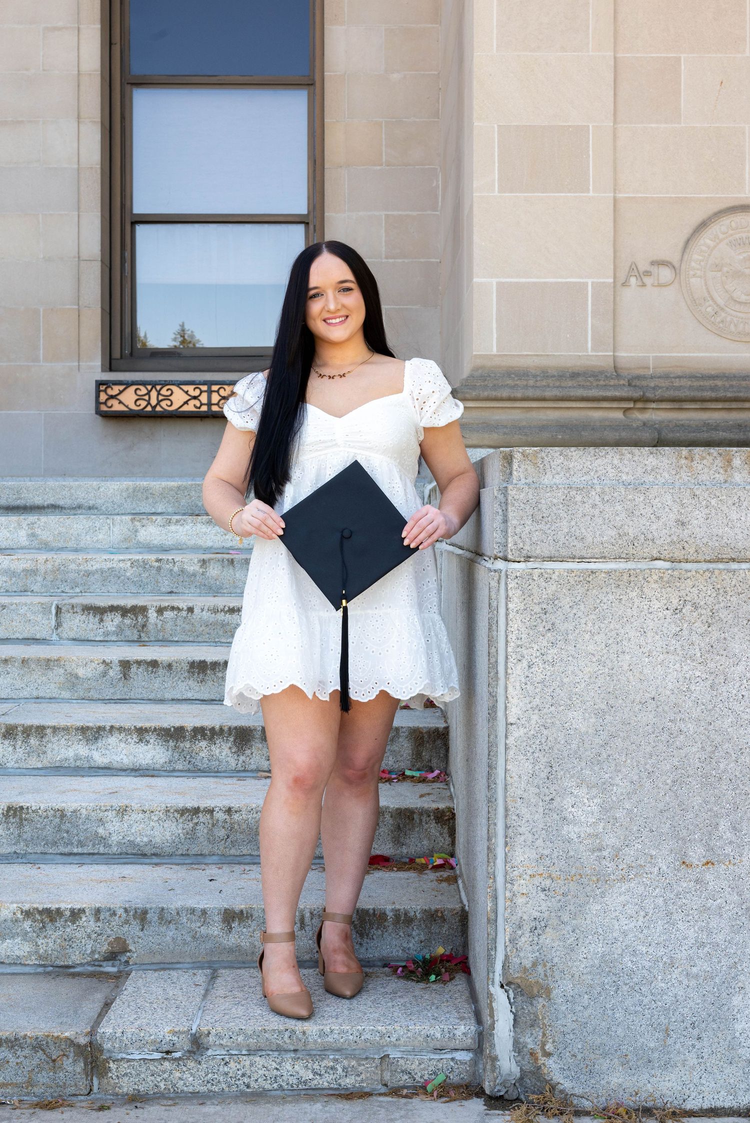 Young woman in white dress holding graduation cap on stone steps of a university building.
