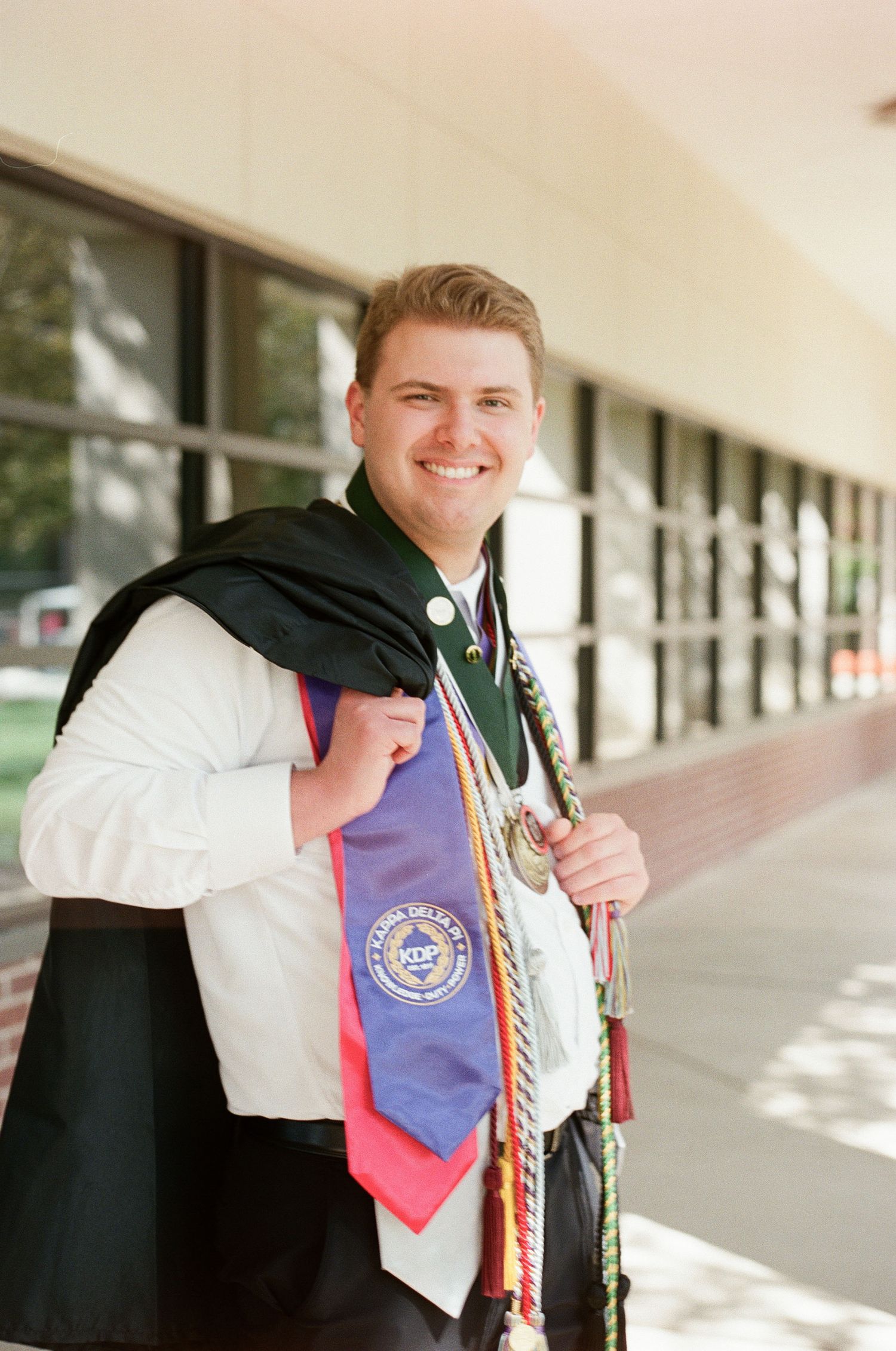 Smiling male graduate wearing green academic hood and colorful honor cords and stoles outside a university building.
