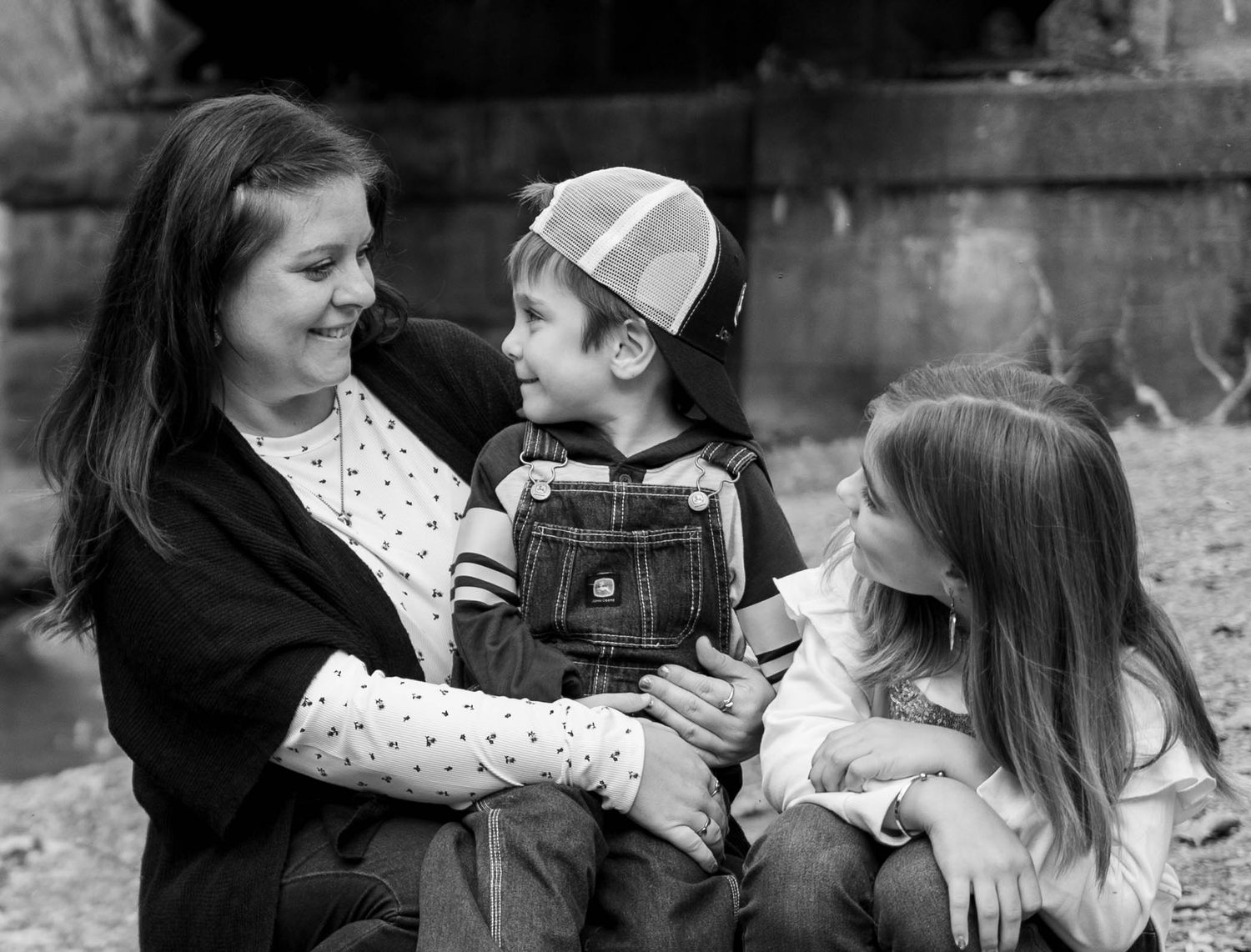 Black and white photo of a smiling woman holding a young boy in overalls and cap, with a girl beside them outdoors.