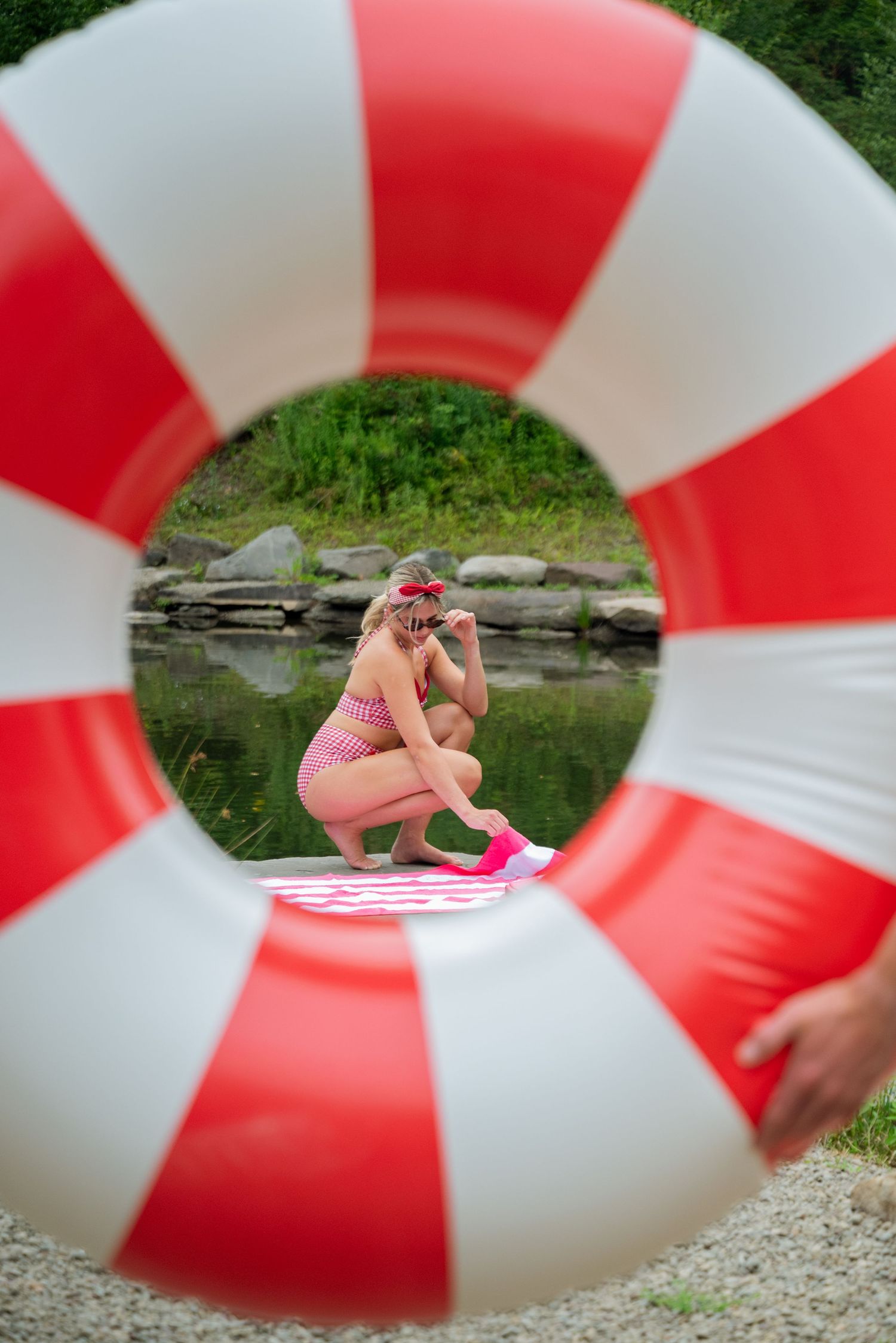 Girl in pink bikini crouching by a pond, viewed through a red and white striped inflatable ring float.