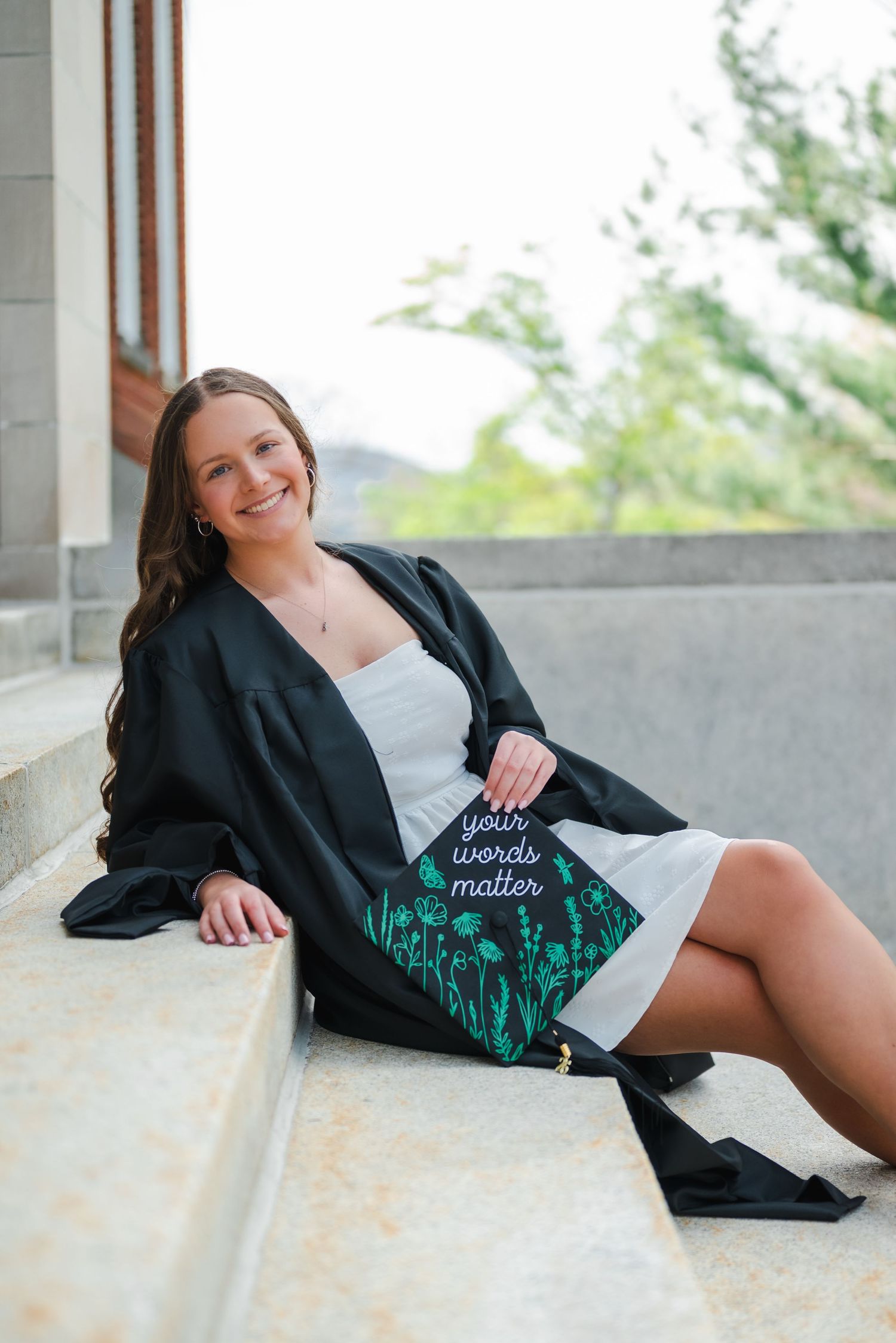 Smiling graduate in black gown sits on steps holding a teal mortarboard cap reading your words matter.