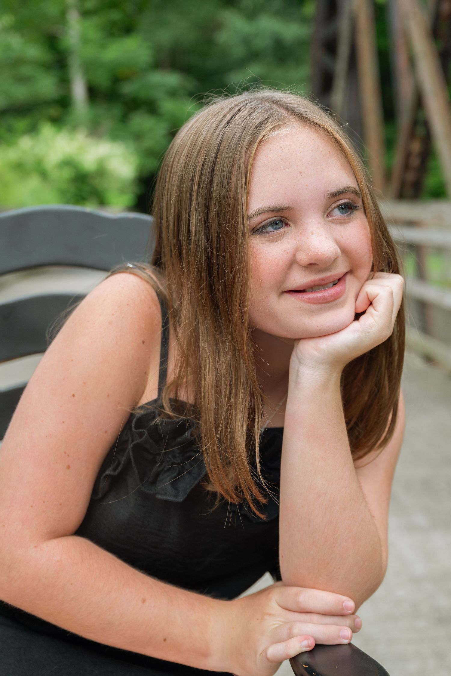 A smiling teenage girl with long brown hair rests her chin on her hand while sitting on a bench outdoors.