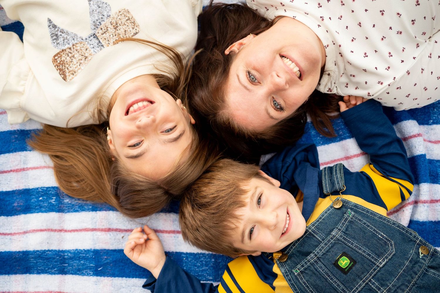 Three happy children lying on striped blanket looking up at camera overhead.
