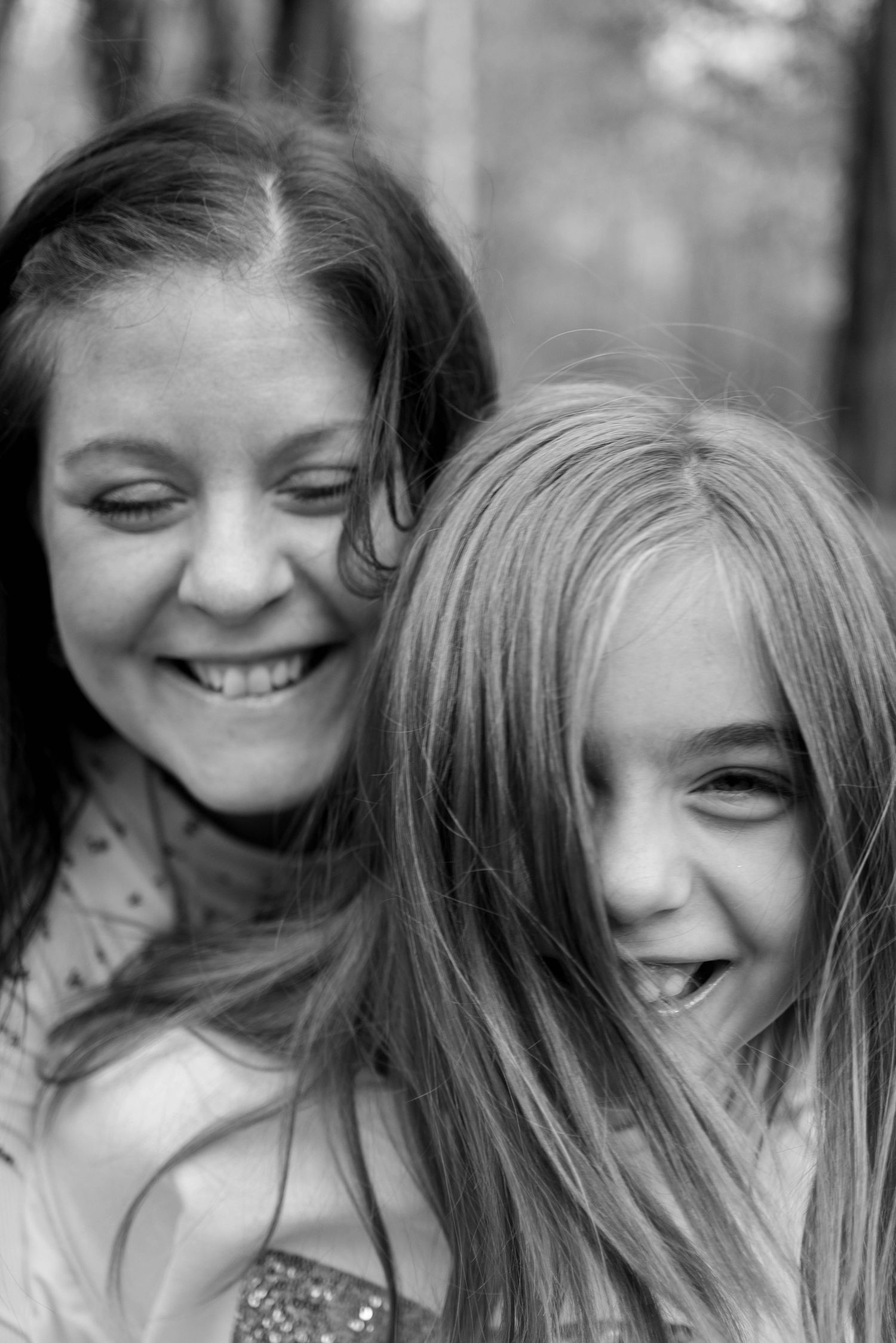 A mother and daughter laugh together in a close-up black and white outdoor portrait among trees.