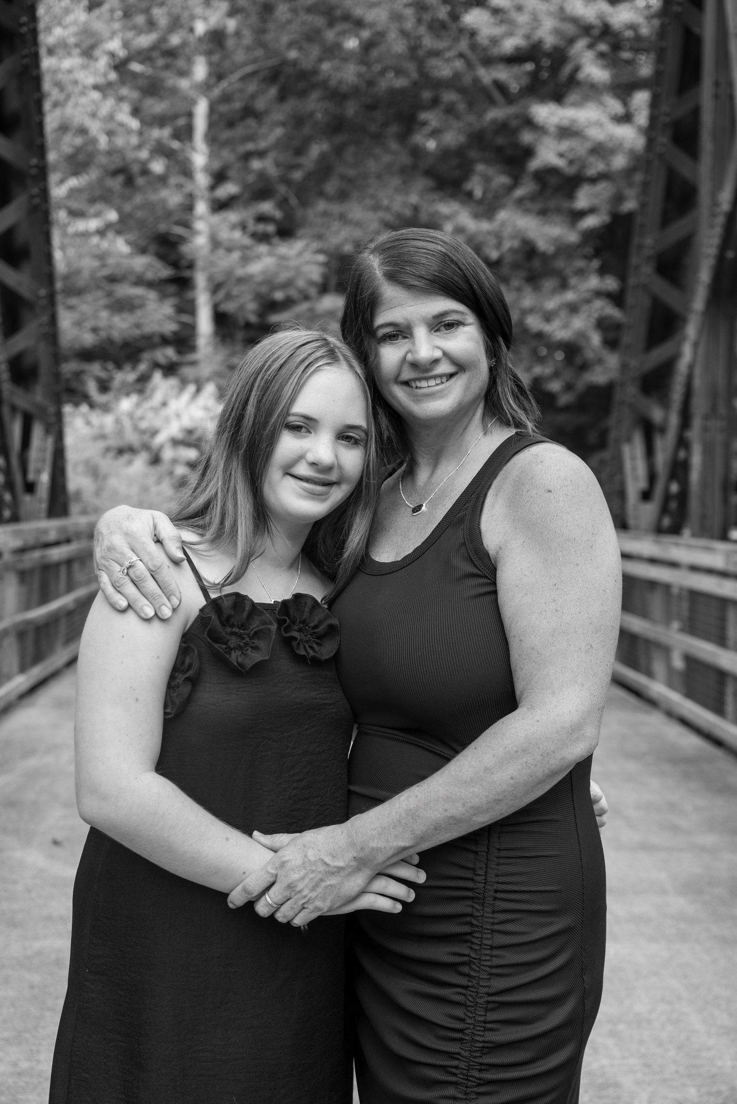 Black and white photo of two women in black dresses smiling on a wooden bridge surrounded by trees.