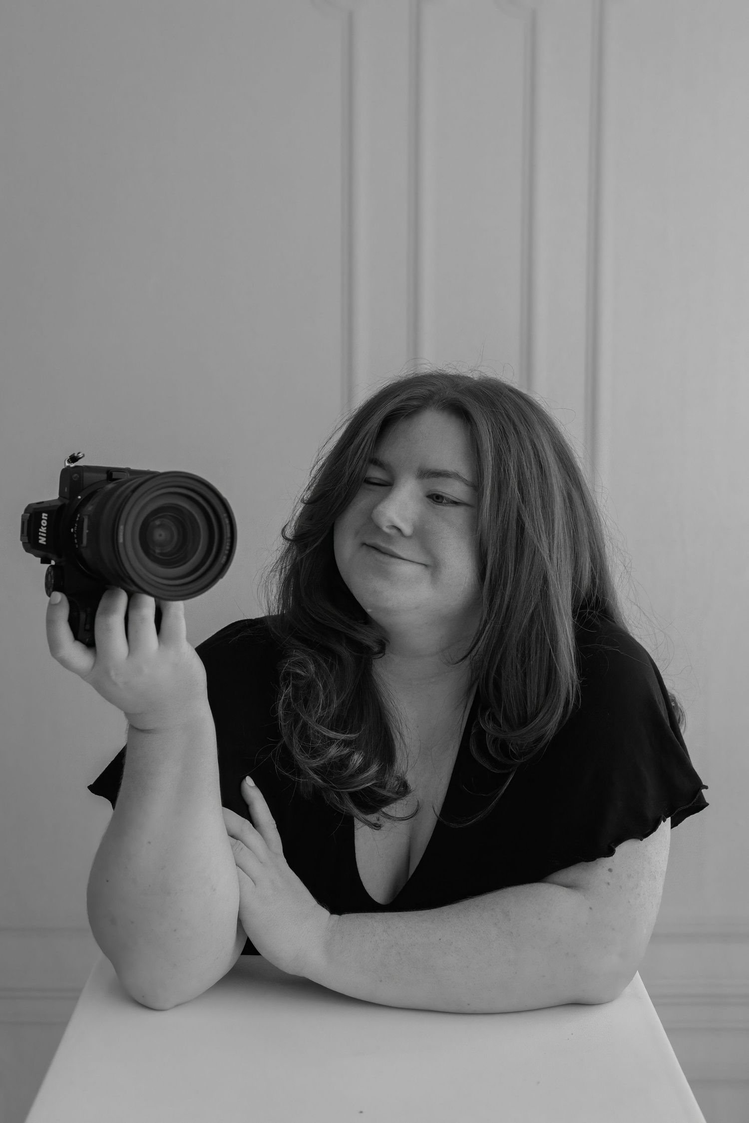 Black and white portrait of a woman holding a Nikon camera, sitting at a white table in a bright studio.