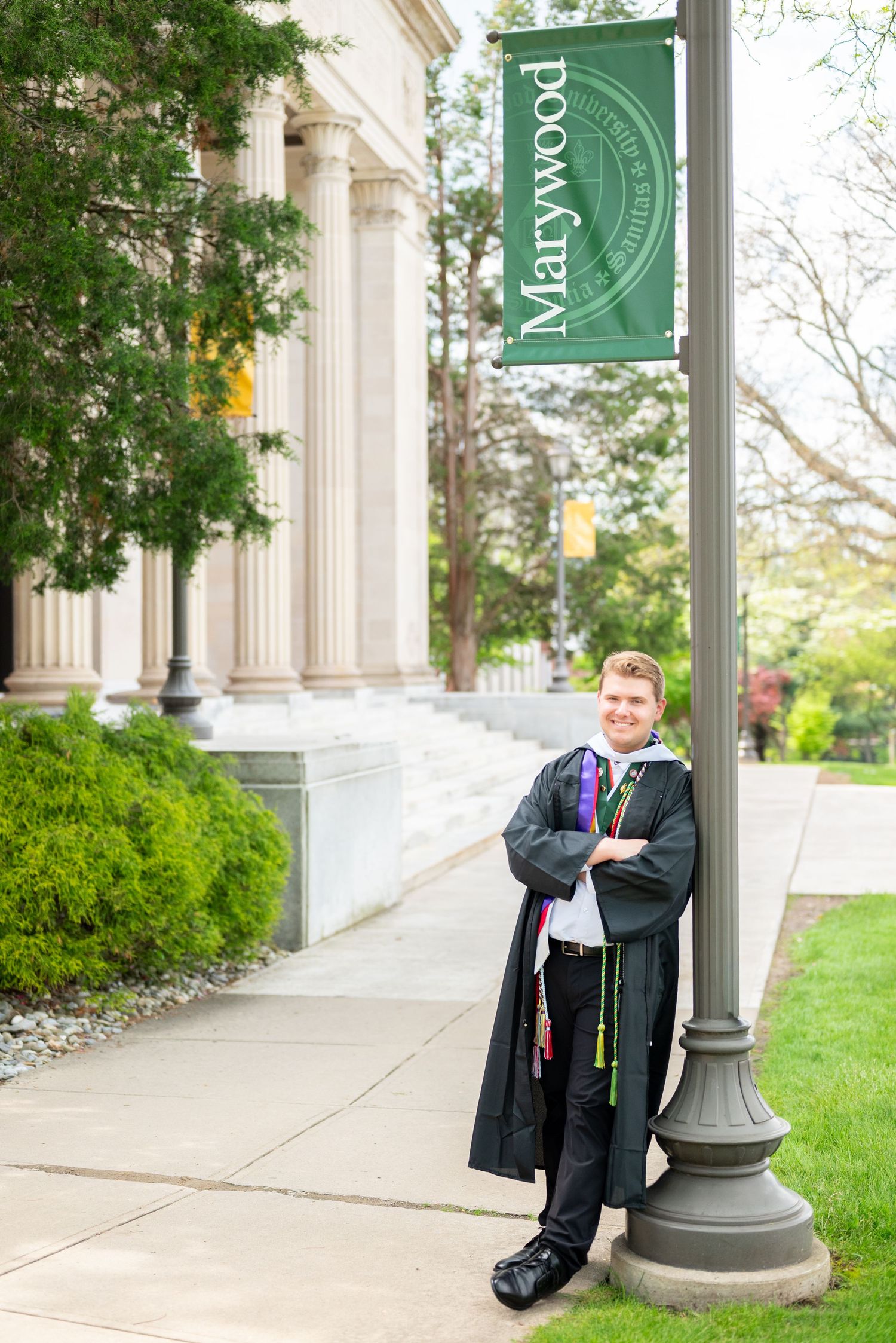 Graduate in cap and gown leaning against a Marywood University banner pole on campus sidewalk.