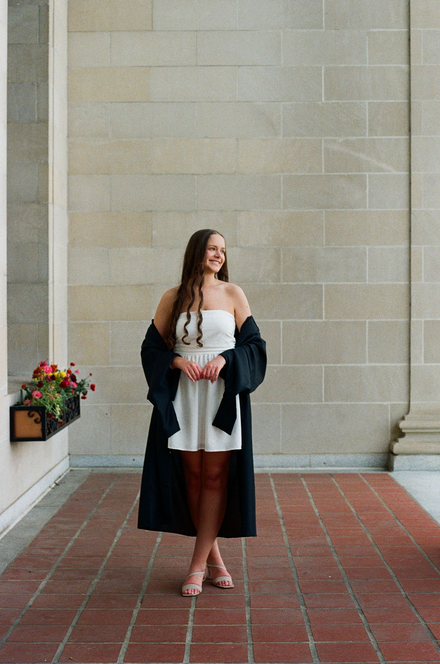Young woman in white dress and black graduation gown smiles outdoors near stone columns on brick floor.