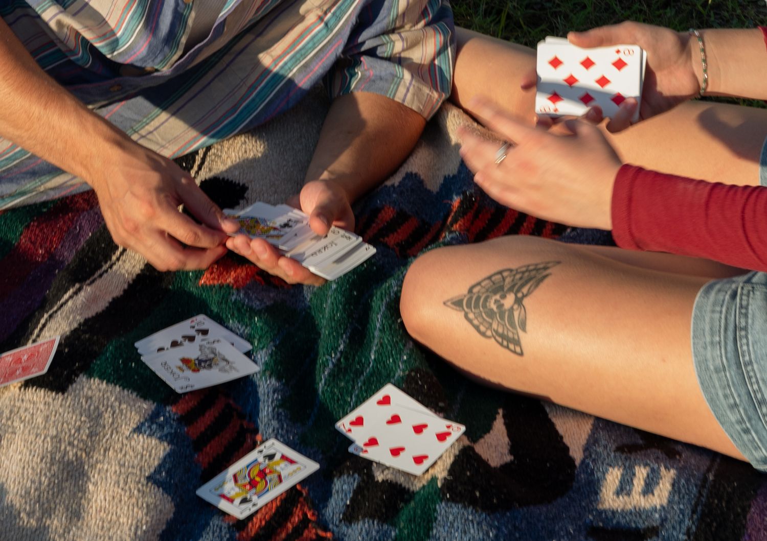 Two people playing cards outdoors on a blanket, with tattoo visible on one person's knee.