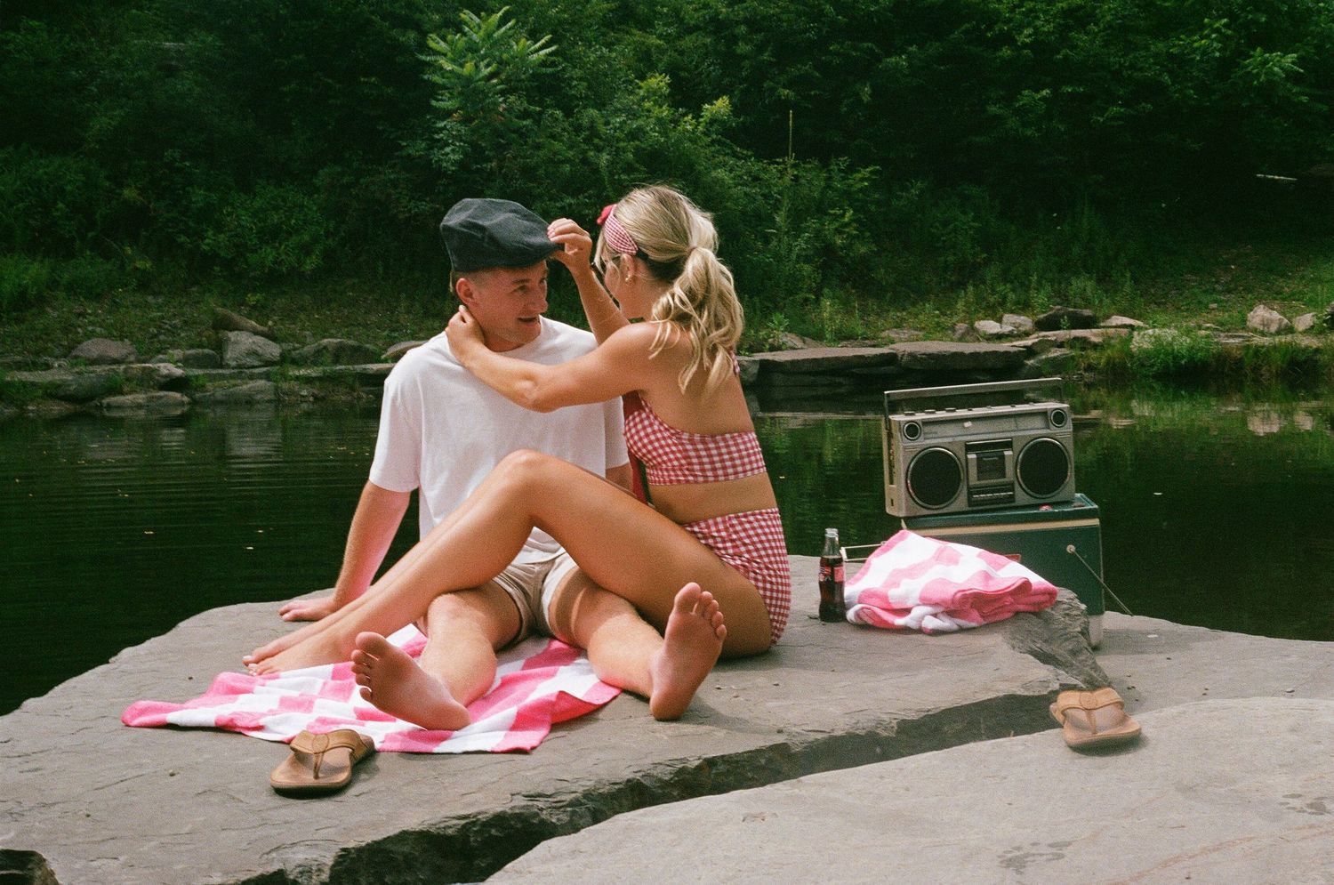Couple relaxing on a rock by a lake with a boombox, sharing a romantic picnic moment outdoors.