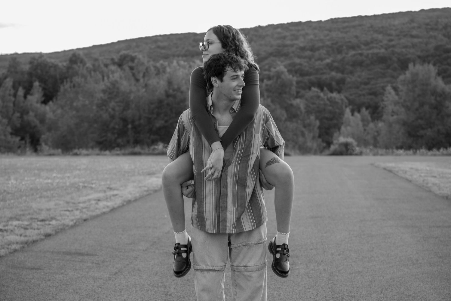 Black and white photo of a young man giving a woman a piggyback ride on an open road surrounded by trees.