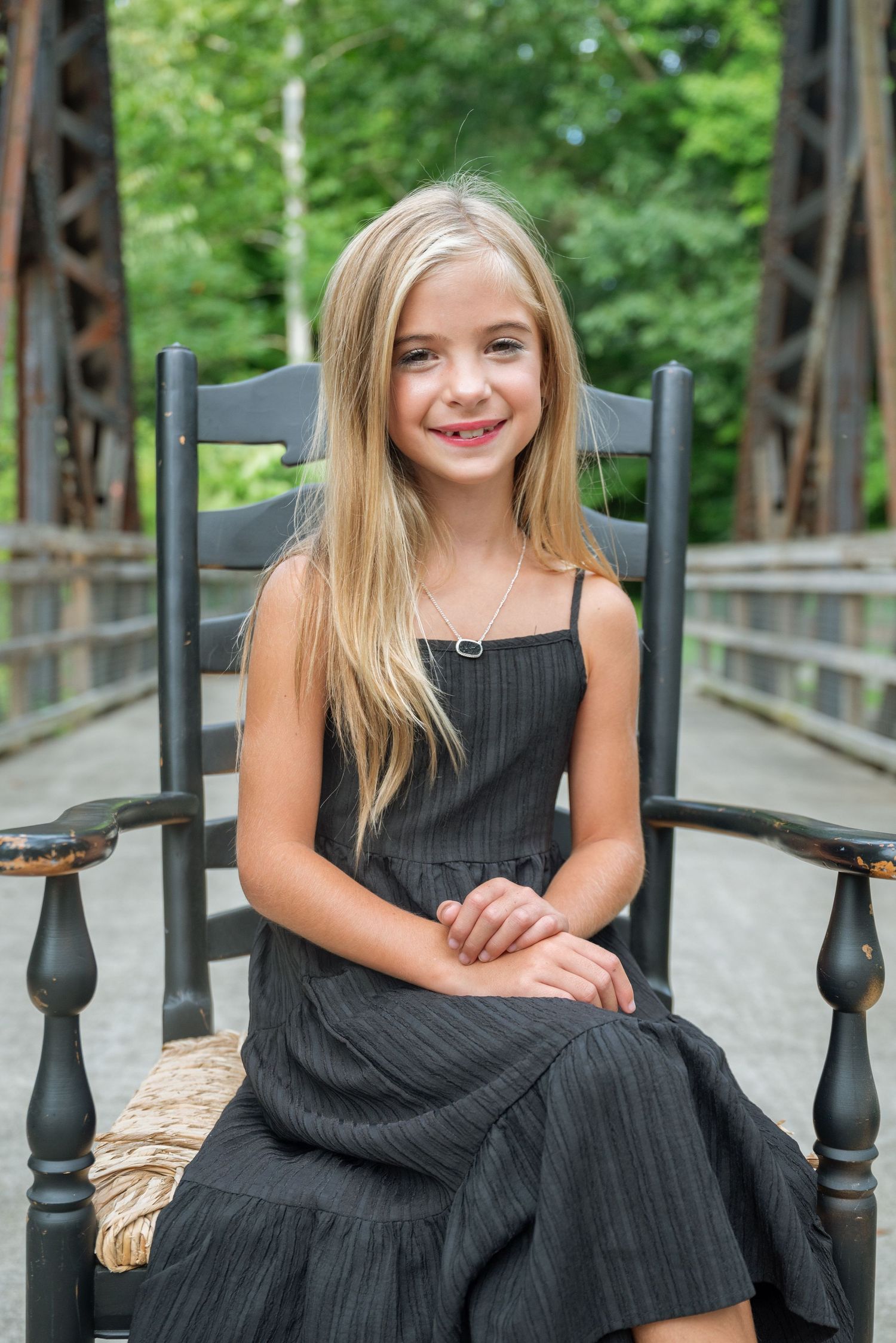 Young girl with long blonde hair sitting in black rocking chair on bridge wearing dark dress.