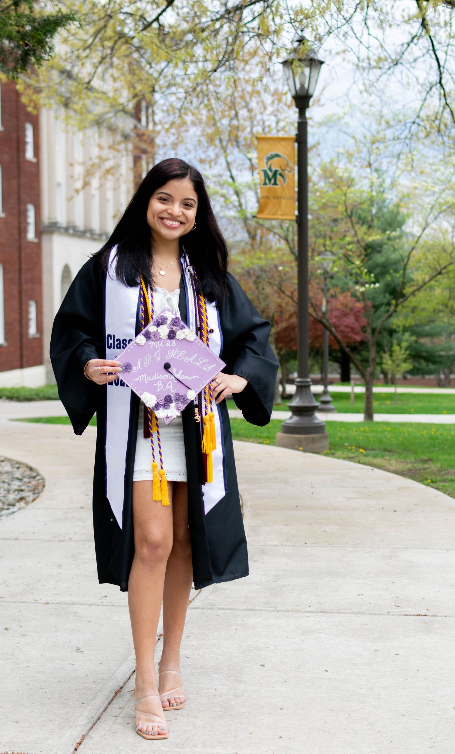 Smiling graduate in cap and gown holding decorated mortarboard on college campus.