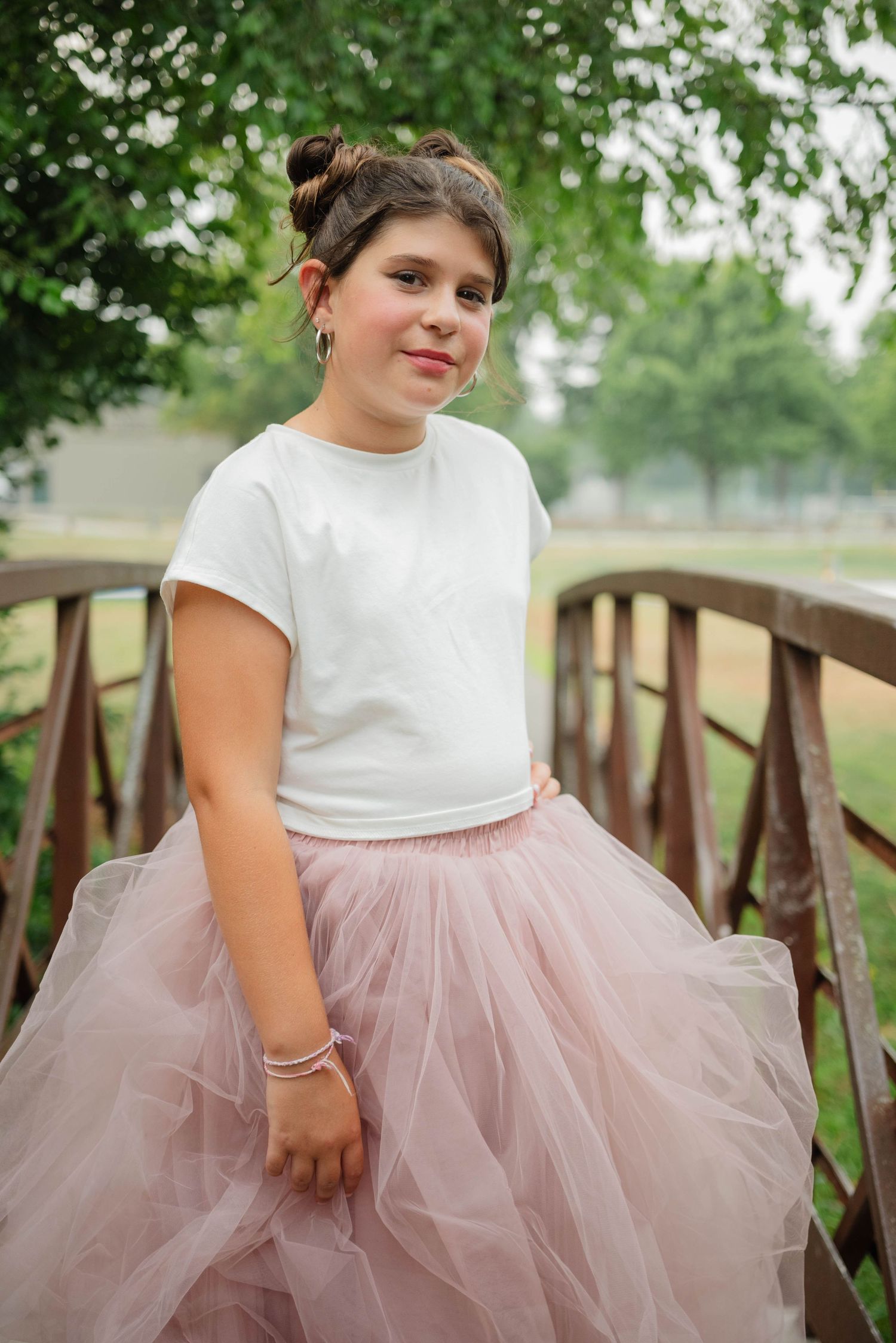 Young girl in white tee and pink tulle skirt posing on a wooden bridge with green trees behind.