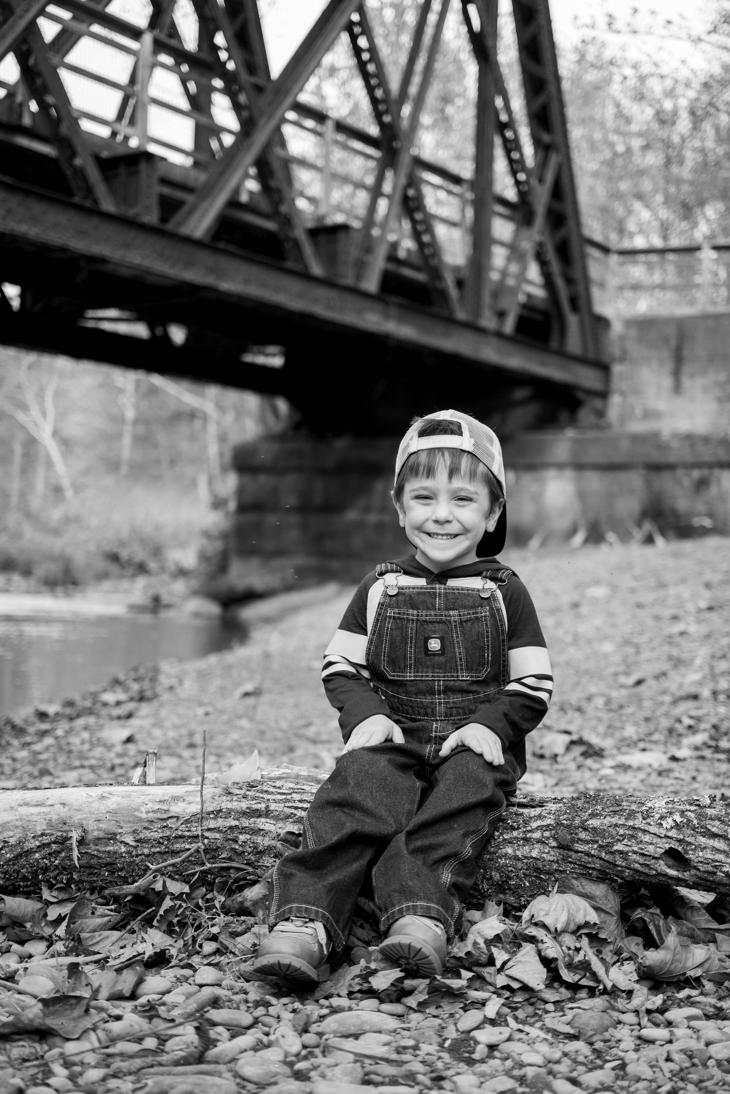 Smiling young boy in overalls and backwards cap sits on a log near a bridge in black and white photo.