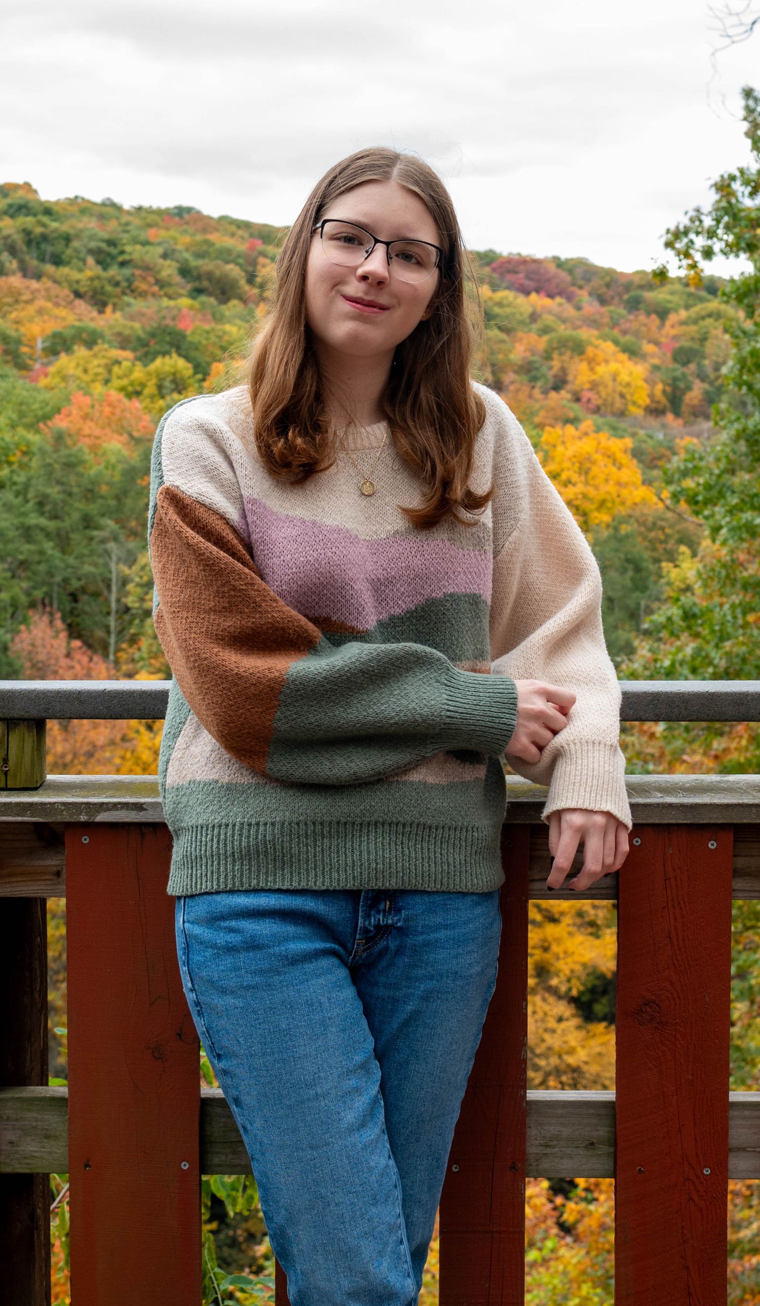 Teen girl in colorblock sweater and jeans leans on wooden deck railing with fall foliage backdrop.