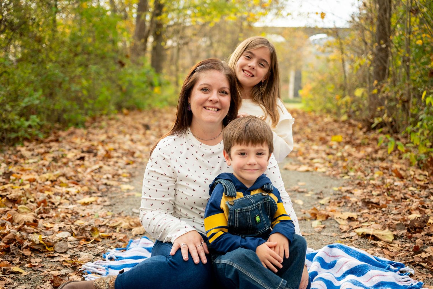 A mother sits on a blanket with her two children smiling on an autumn leaf-covered path.