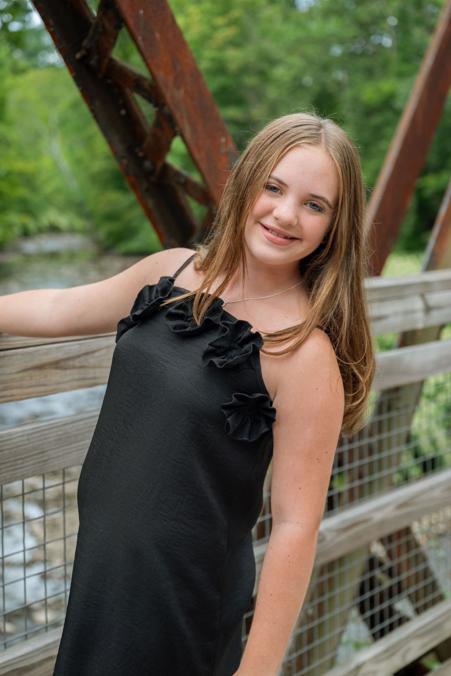 A smiling young girl in a black ruffle dress poses on a wooden bridge surrounded by lush green trees.