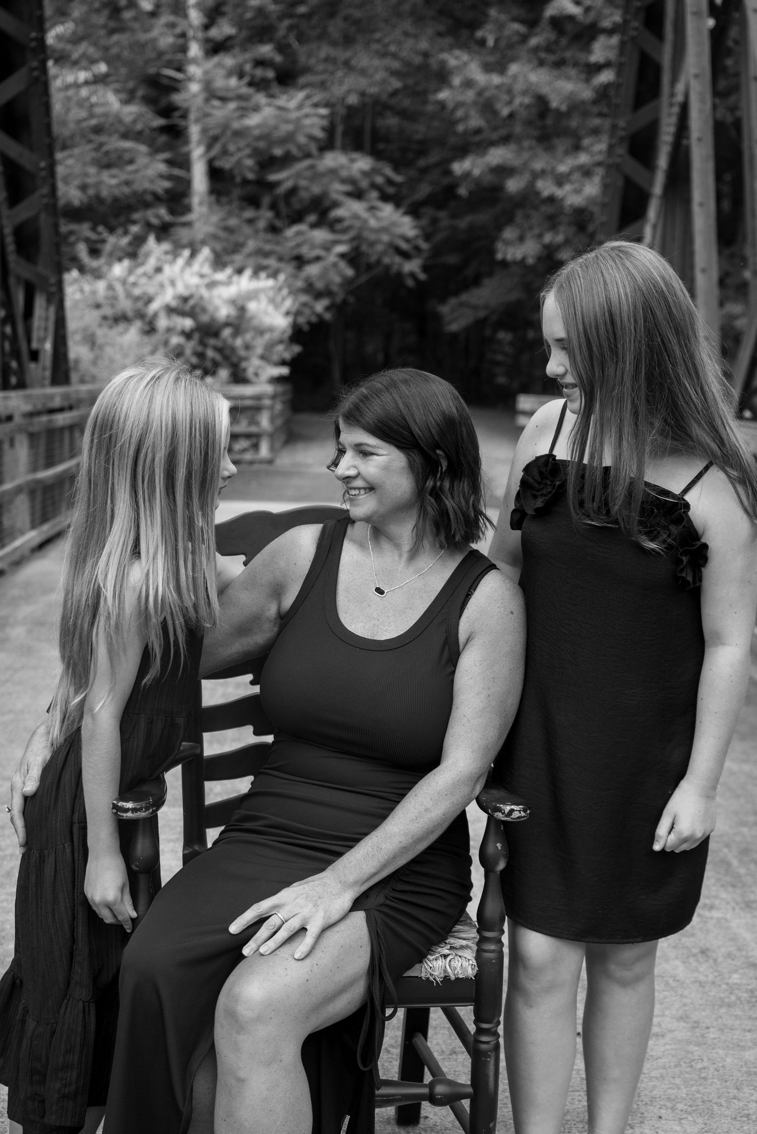 Black and white photo of a woman seated on a chair smiling at two blonde girls in black dresses on a bridge.