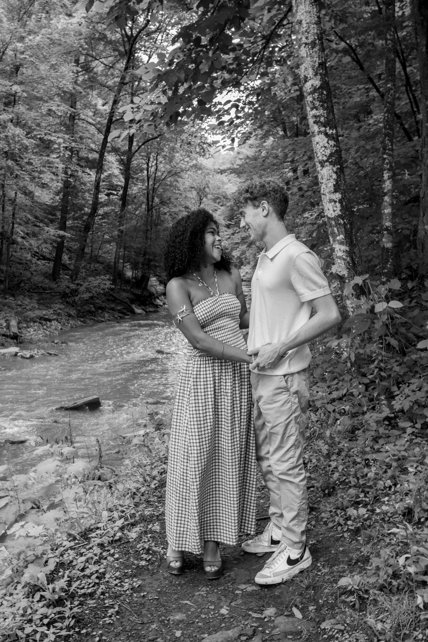 Black and white photo of a couple holding hands and smiling at each other beside a forest stream.