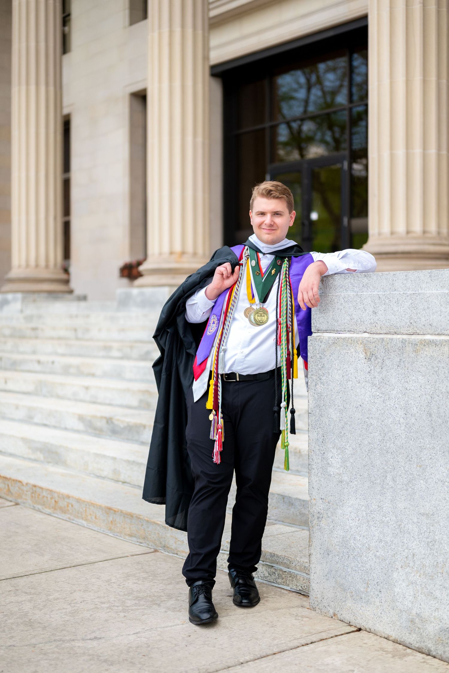 Young graduate poses outdoors in academic regalia with colorful honor cords and medal near stone columns.