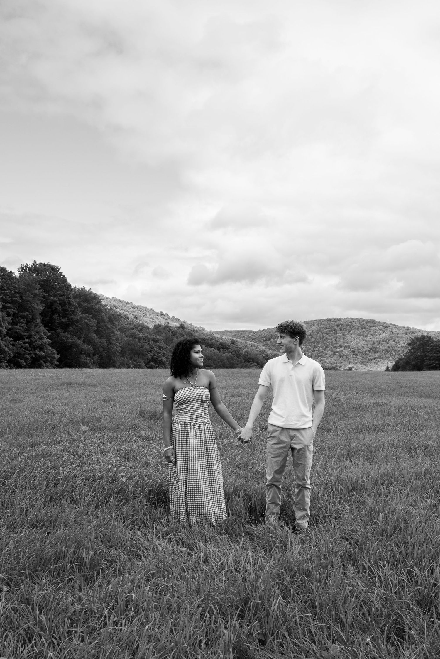 Black and white photo of a couple holding hands in a grassy field with rolling hills and trees in the background.