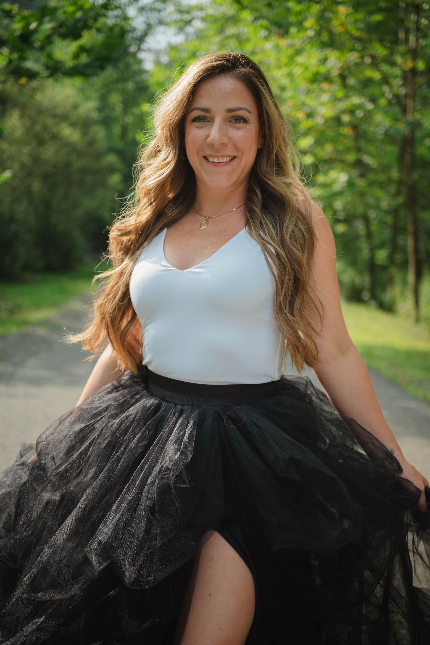 Smiling woman with long wavy hair wearing white top and black tulle skirt on a park road.