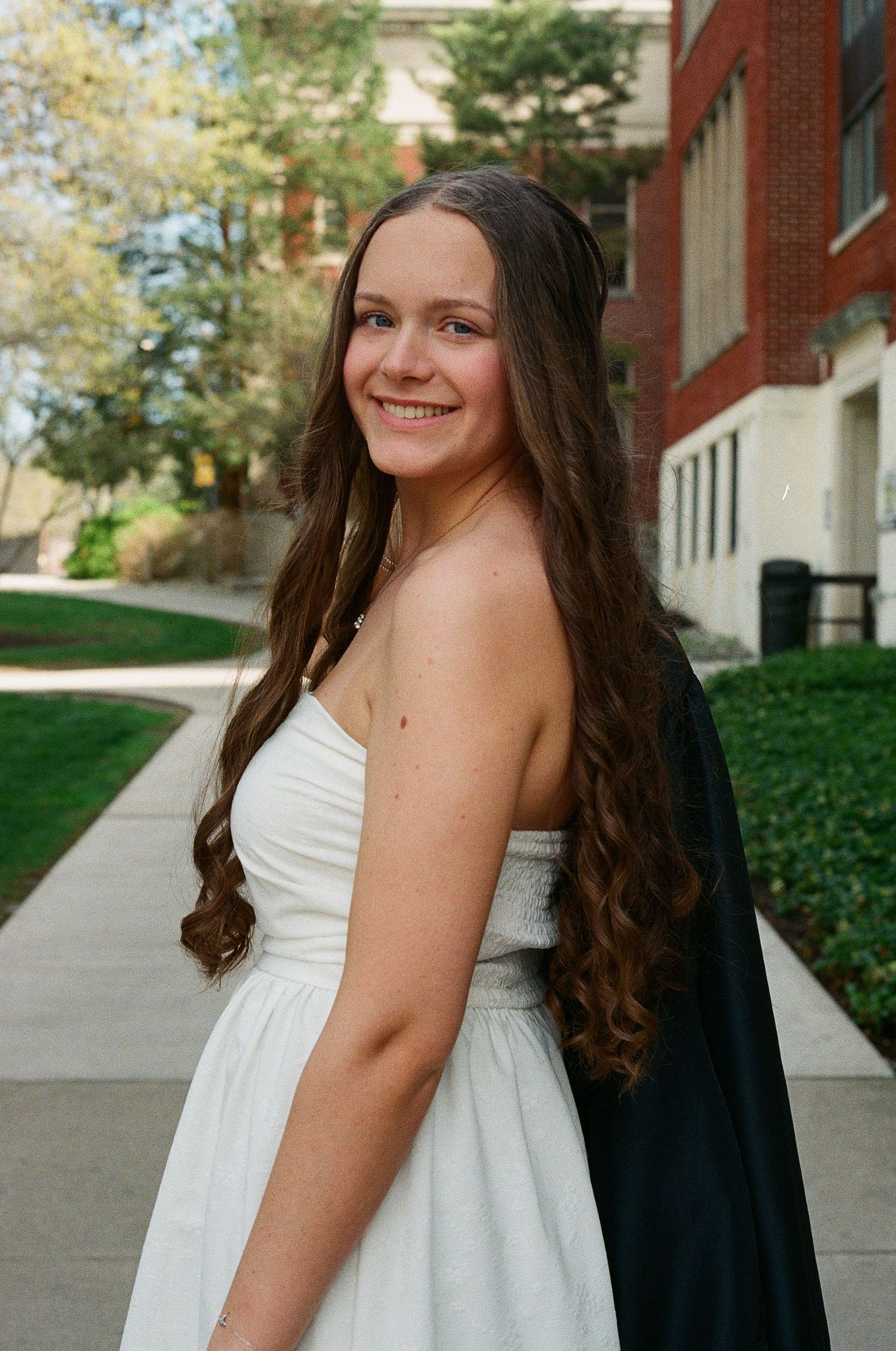 Smiling young woman with long brown hair wearing a white strapless dress outdoors near a brick building.
