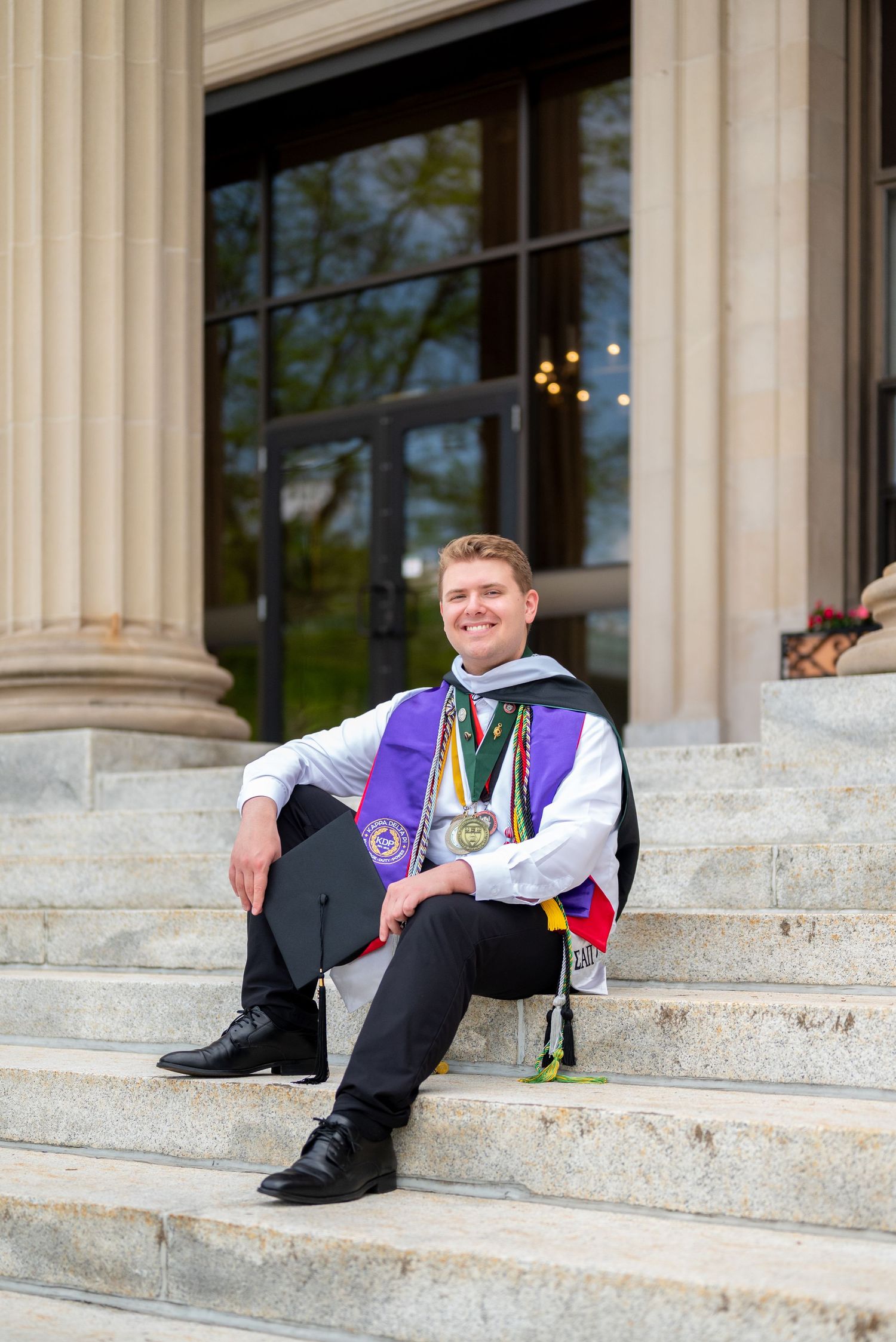 Smiling college graduate sitting on stone steps wearing purple sash, medals, and holding cap in graduation attire.