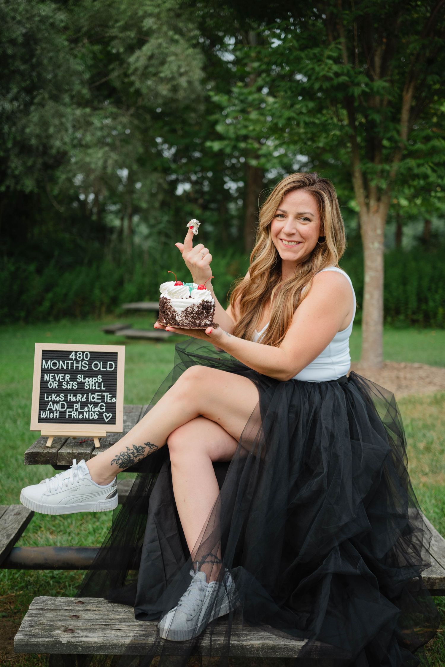 Smiling woman in black tulle skirt holding cake on picnic bench with 480 months old sign outdoors.