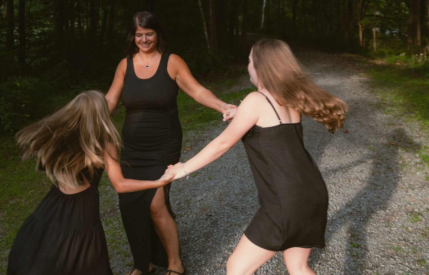 A woman in a black dress holds hands with two girls in black dresses, spinning together on a gravel path in a forest.