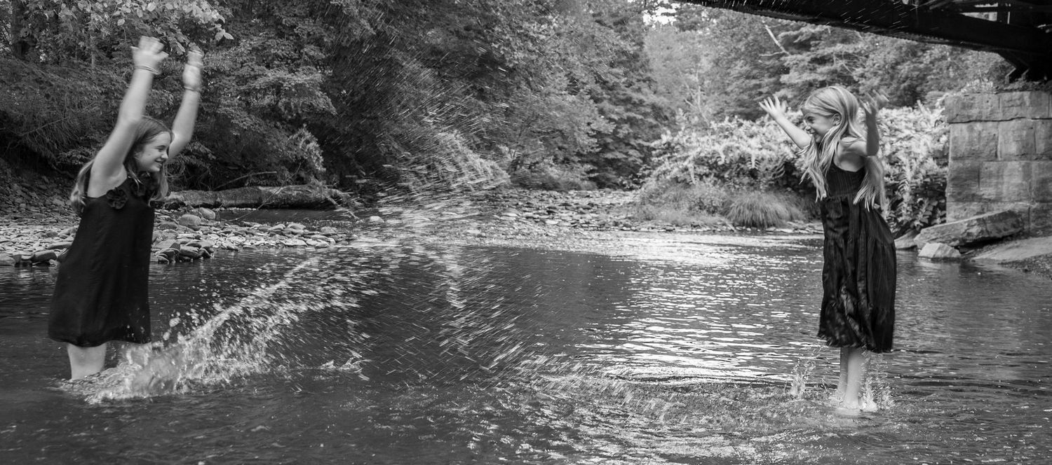 Two girls in black dresses splashing water in a shallow creek surrounded by lush greenery.