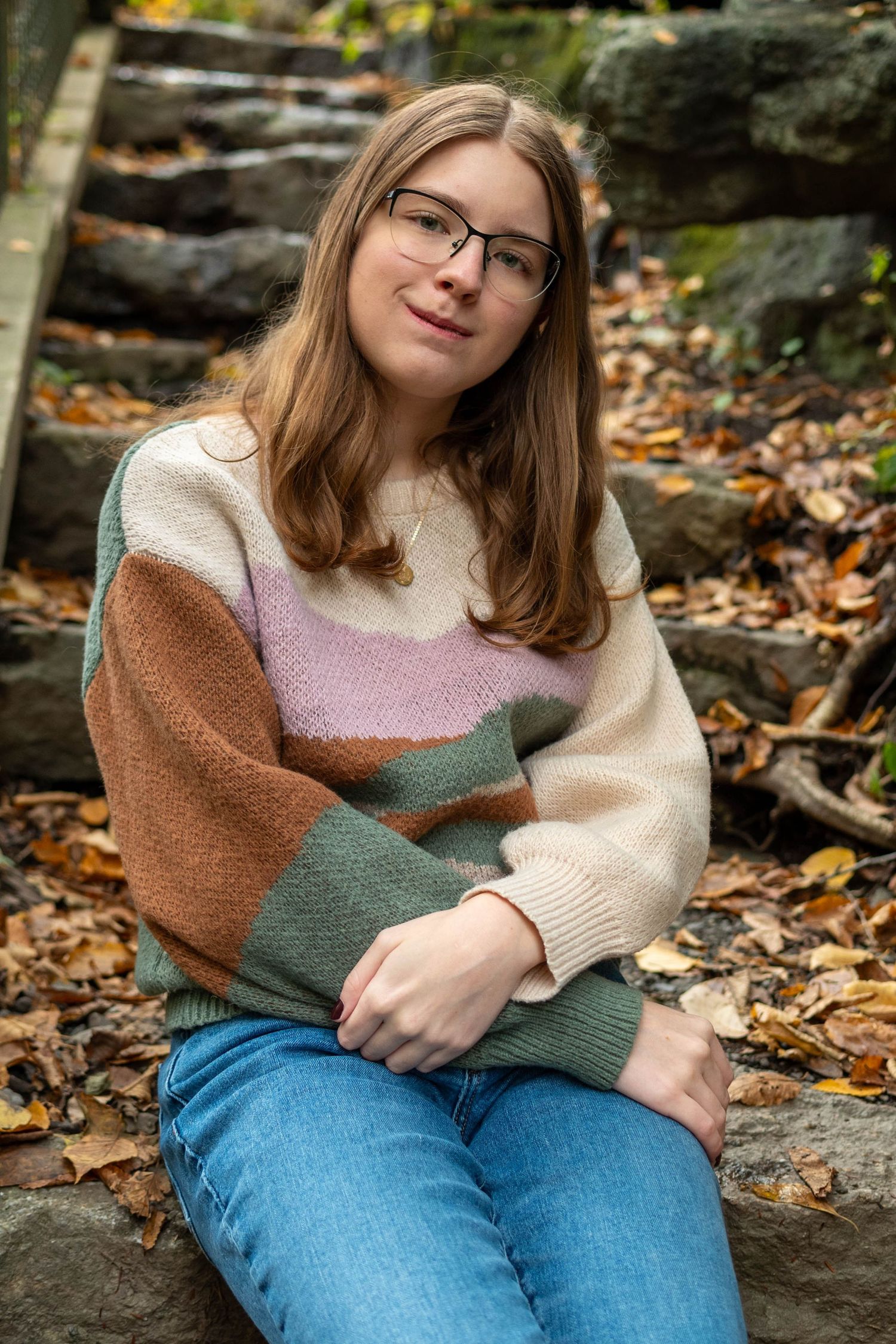 Teen girl with glasses in colorblock sweater sits on stone steps surrounded by fall foliage.