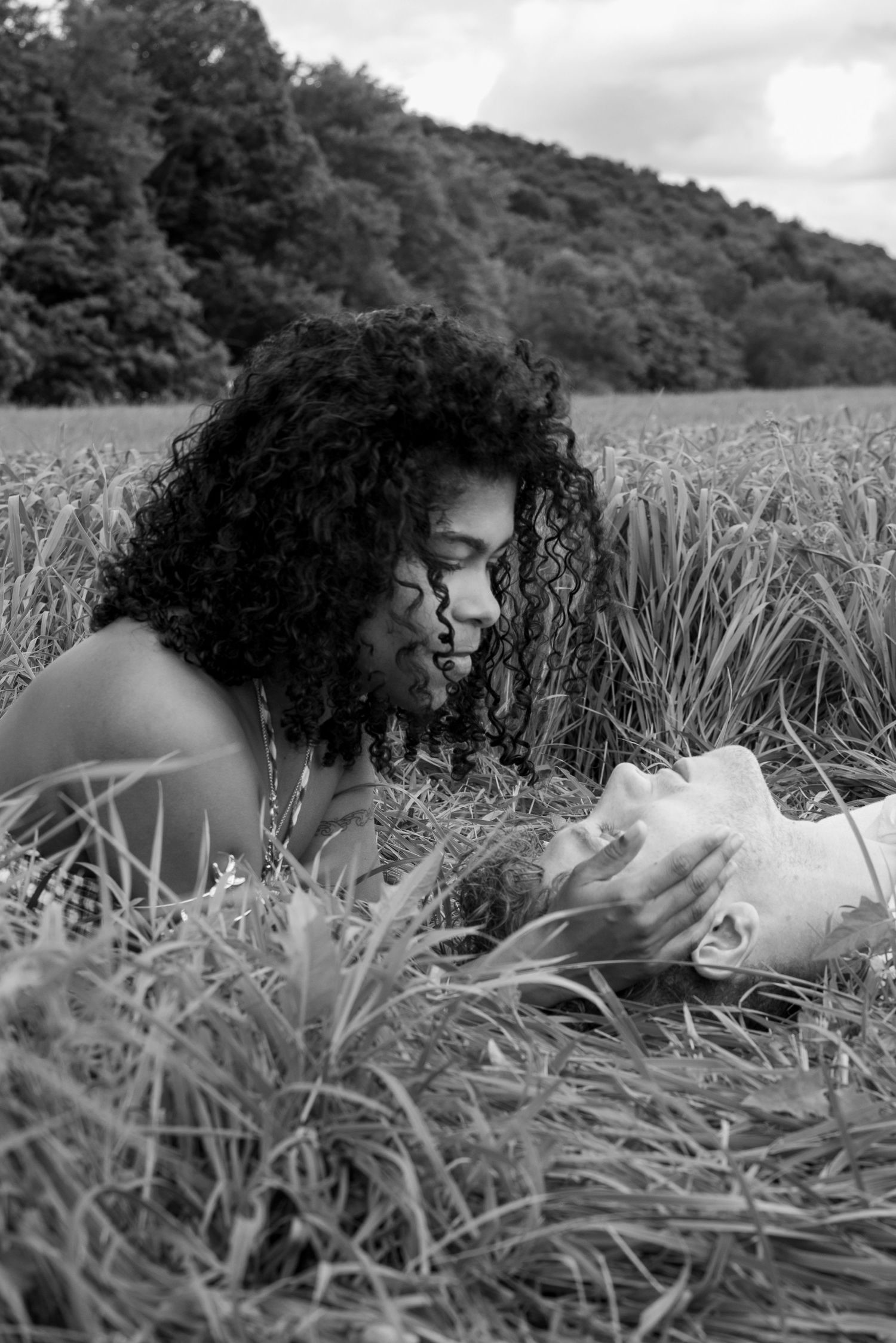Black and white photo of a woman with curly hair lying in a field, gazing at a seashell among tall grass.