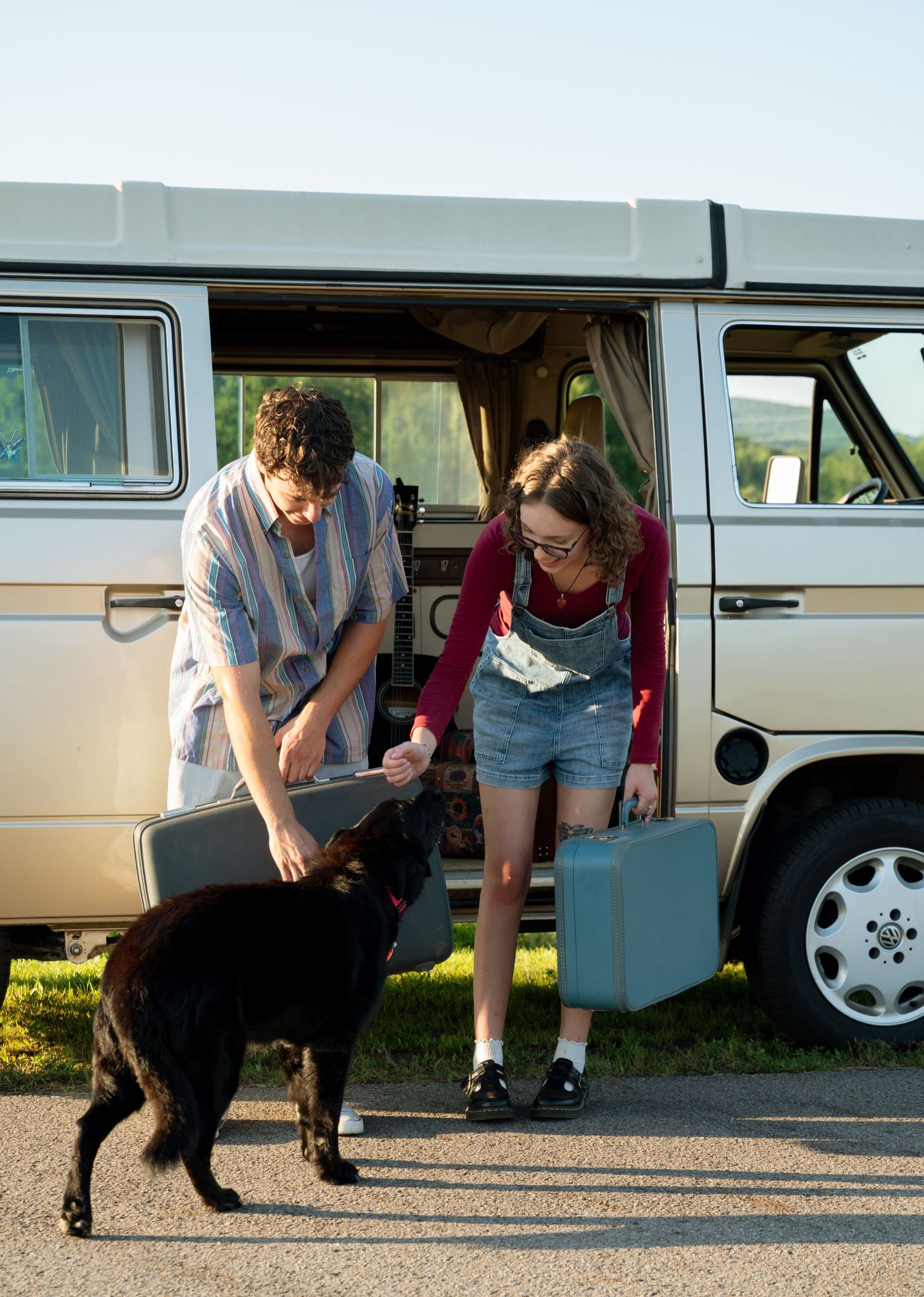 A couple with a black dog unloading luggage from a vintage VW van on a sunny day.