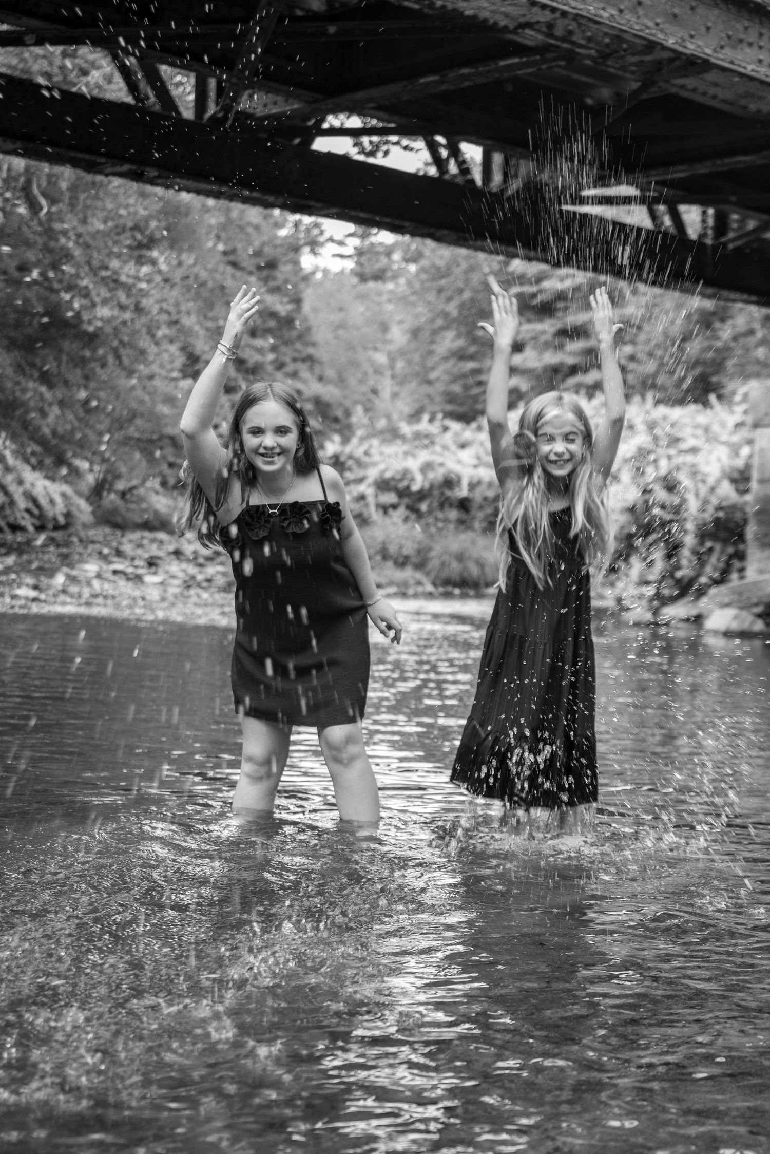 Two girls in black dresses joyfully splash in a river under a bridge, arms raised, in a black and white photo.