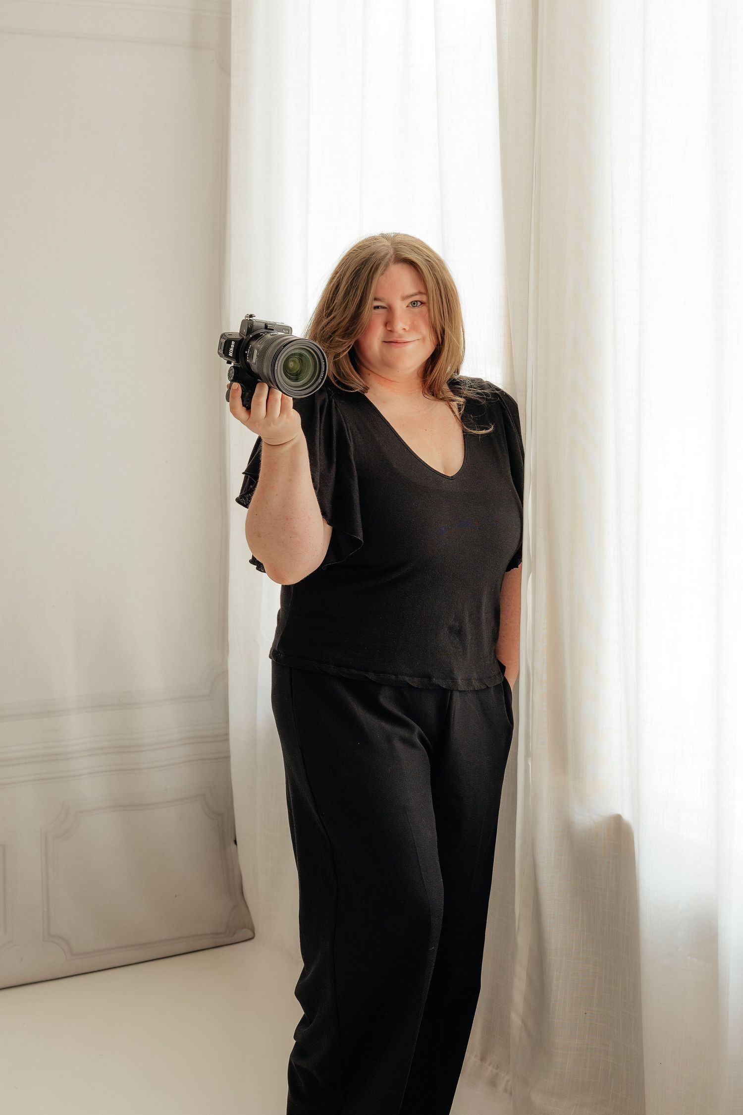 A smiling female photographer in black outfit holds a professional camera while standing near white curtains in a bright studio.