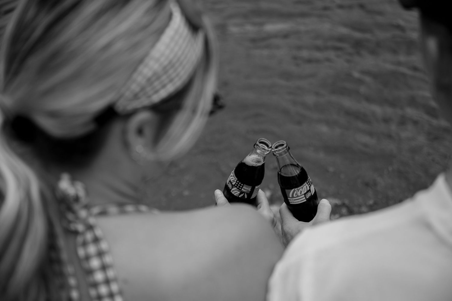 Two people clinking Coca-Cola bottles together outdoors near water in a black and white photo.