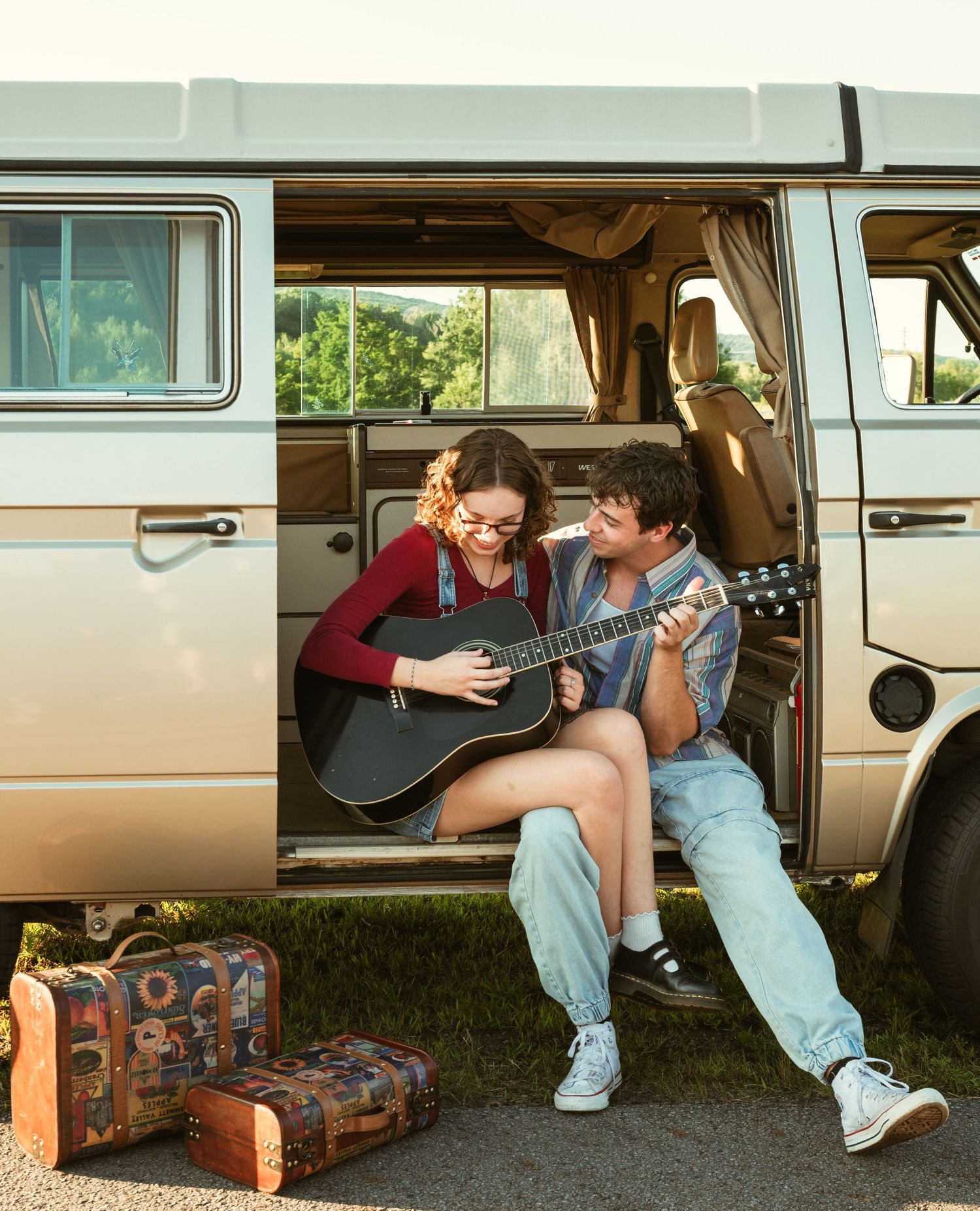 Couple sitting in open van doors playing acoustic guitar with vintage suitcases nearby outdoors.