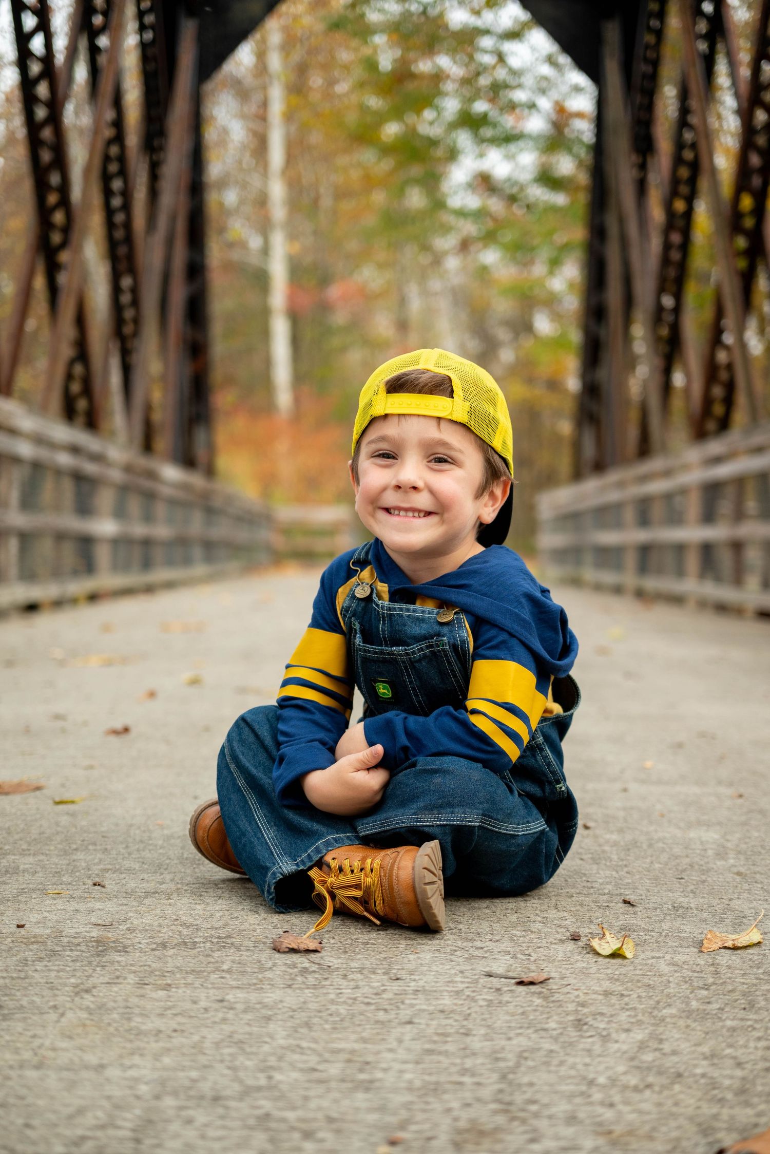 Happy young boy in yellow cap and overalls sits cross-legged on a bridge smiling at the camera.