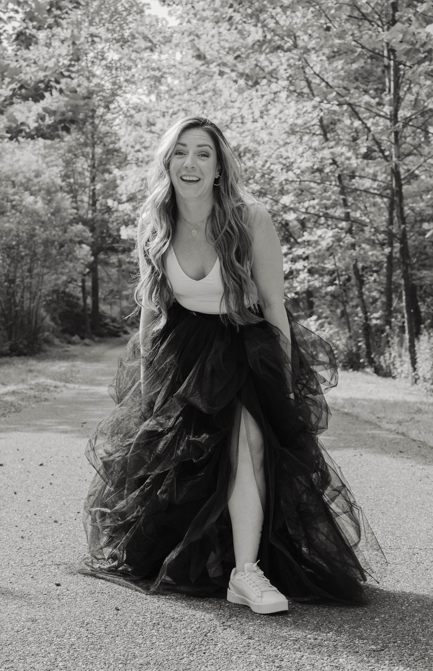 Black and white photo of laughing woman in black tulle skirt and white top walking on wooded path.
