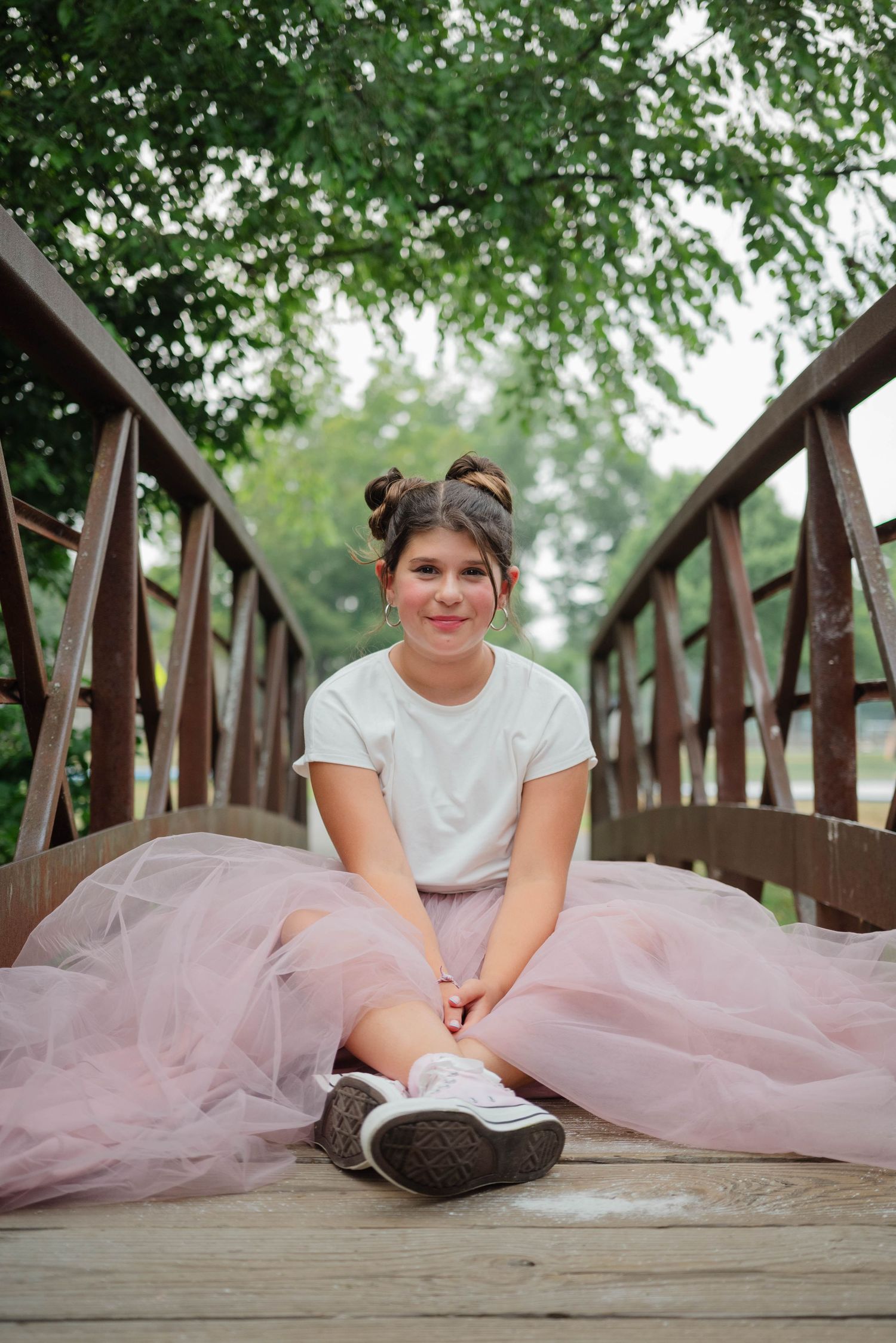 Young girl in pink tulle skirt and white tee sits smiling on wooden bridge surrounded by green trees.