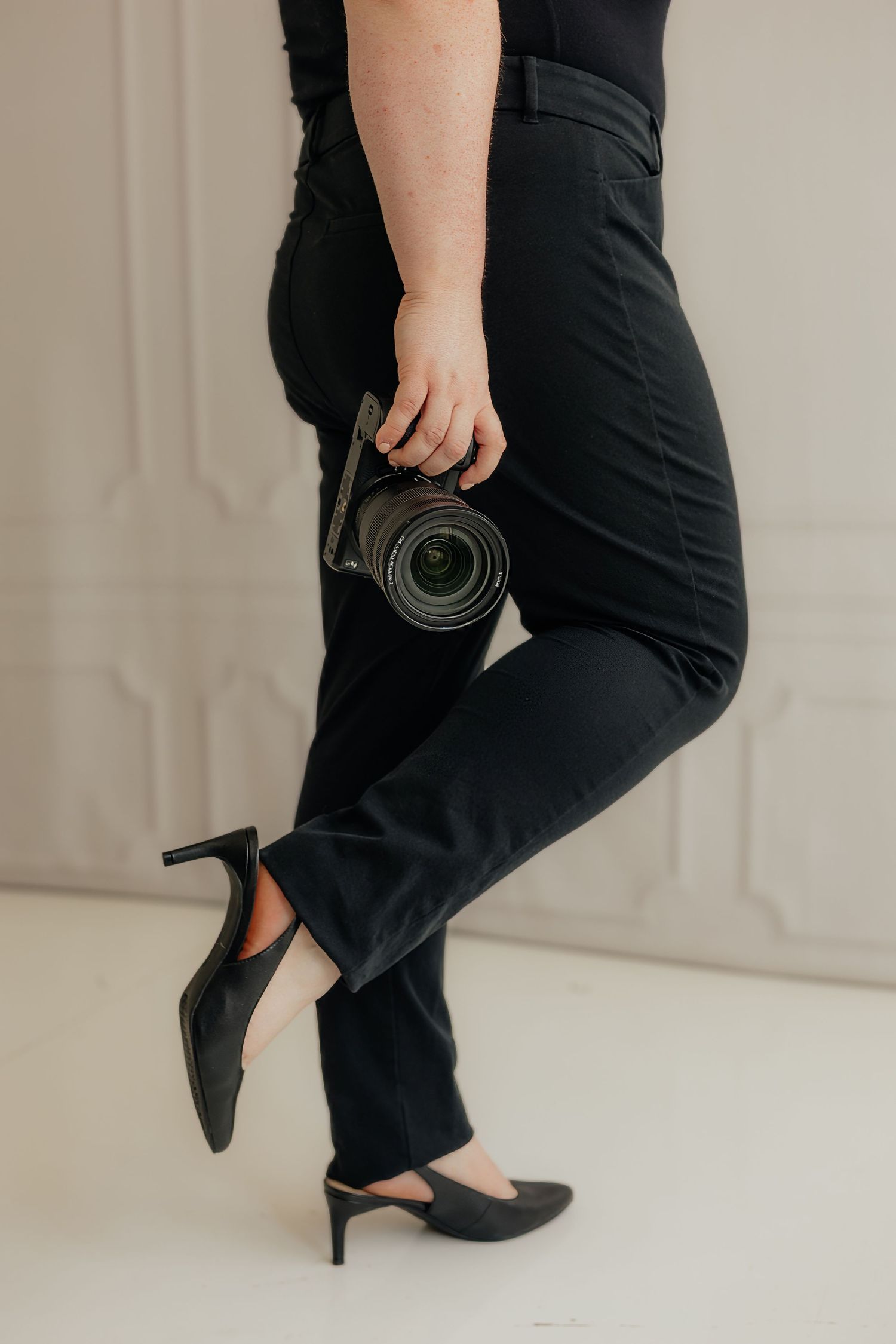 Woman in black outfit and heels holding a DSLR camera, standing against a white decorative wall.